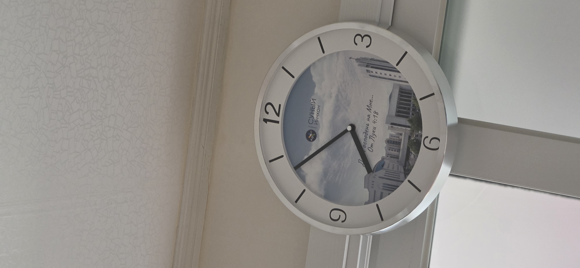 The image is a close-up, eye-level shot of a round wall clock with a picture of the Incheon Catholic Cathedral in Incheon, South Korea, on its face. The clock is white with silver trim, and the numbers 12, 3, 6, and 9 are clearly visible. The hands of the clock are pointing to approximately 11:05. The Incheon Catholic Cathedral, also known as the Cathedral of the Holy Savior of Korea, is depicted in the photograph on the clock face. It is a modern architectural structure with a tall spire. The clock appears to be hanging on a wall near a window or door, as indicated by the bright light and reflections on the right side of the image. The background consists of a textured ceiling and a white wall.