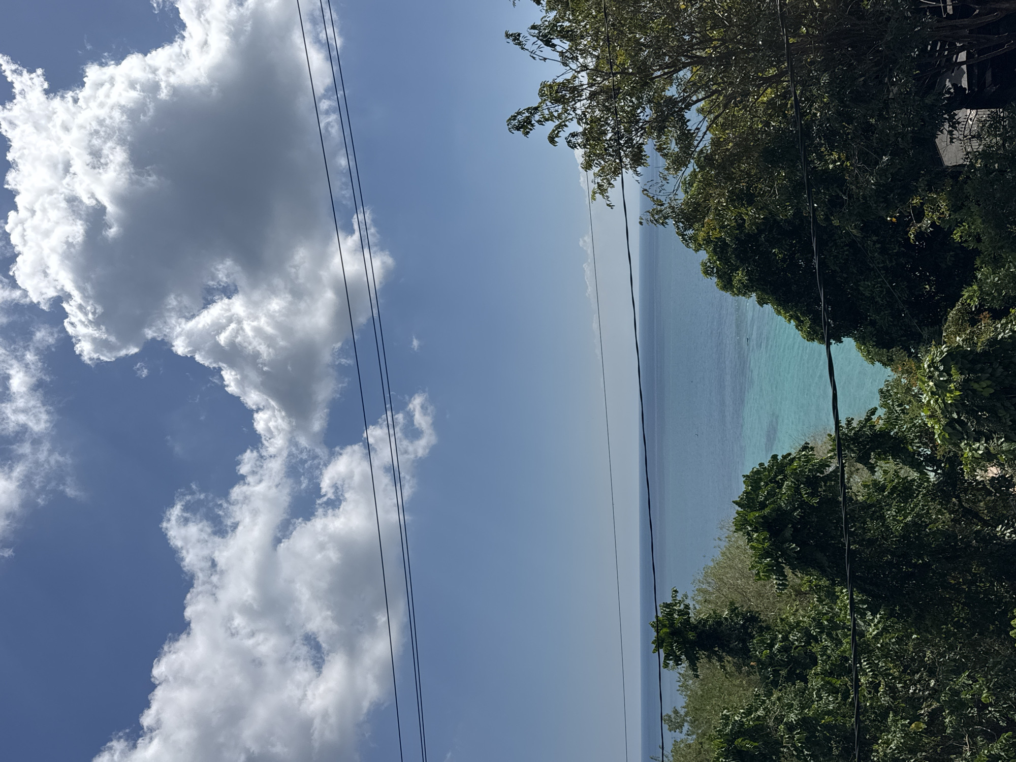 The image depicts a scenic view of the ocean from a slightly elevated position, likely a hillside overlooking the coast. The sky is a vibrant blue, dotted with fluffy white clouds. In the foreground, lush green foliage and trees frame the view, suggesting a tropical or subtropical environment. Power lines are visible across the frame, indicating human infrastructure in the natural landscape. The ocean itself is a striking turquoise color, transitioning to a deeper blue on the horizon. The water appears calm, with a subtle shimmer suggesting sunlight. No people or specific activities are discernible in the image. The overall impression is one of natural beauty and tranquility. This scene is consistent with coastal vistas found in areas like Kuta Selatan, Indonesia, known for its tropical beaches and lush vegetation.