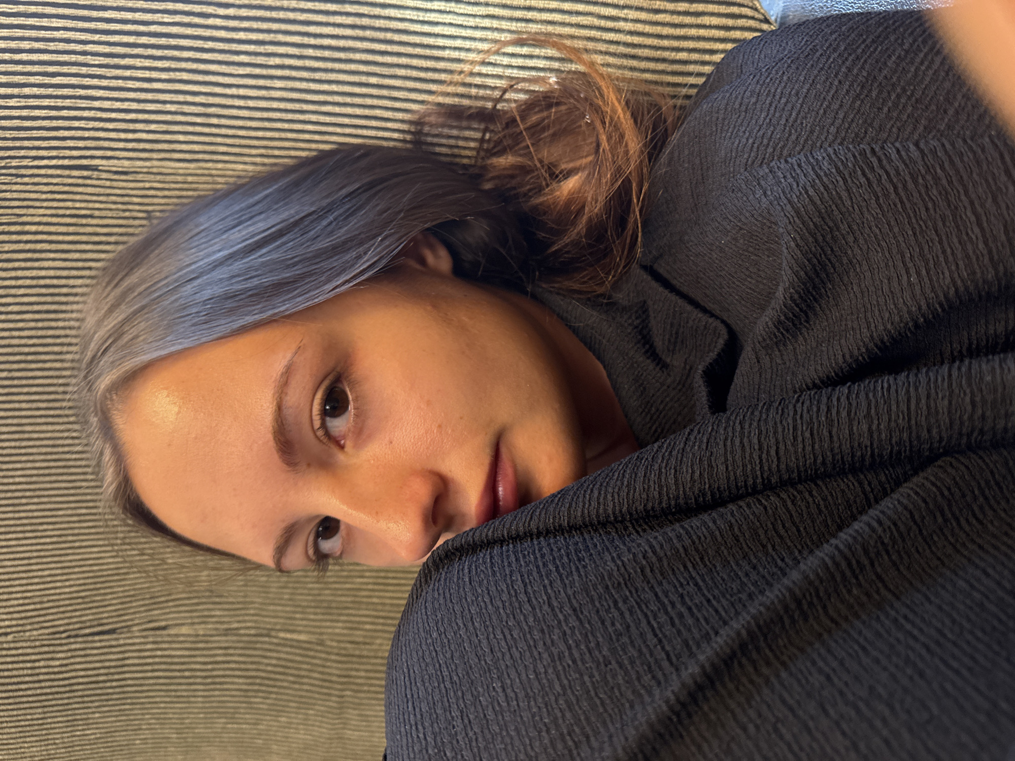 A woman is captured in a close-up shot, her face turned towards the camera with a portion of her shoulder and upper chest obscured by a dark, textured garment. Her hair, a mix of grey and brown hues, is pulled back. She is situated against a backdrop of vertical striped upholstery. The scene appears to be indoors, possibly a domestic setting. There is no indication of the location in Skala Marion, Greece.