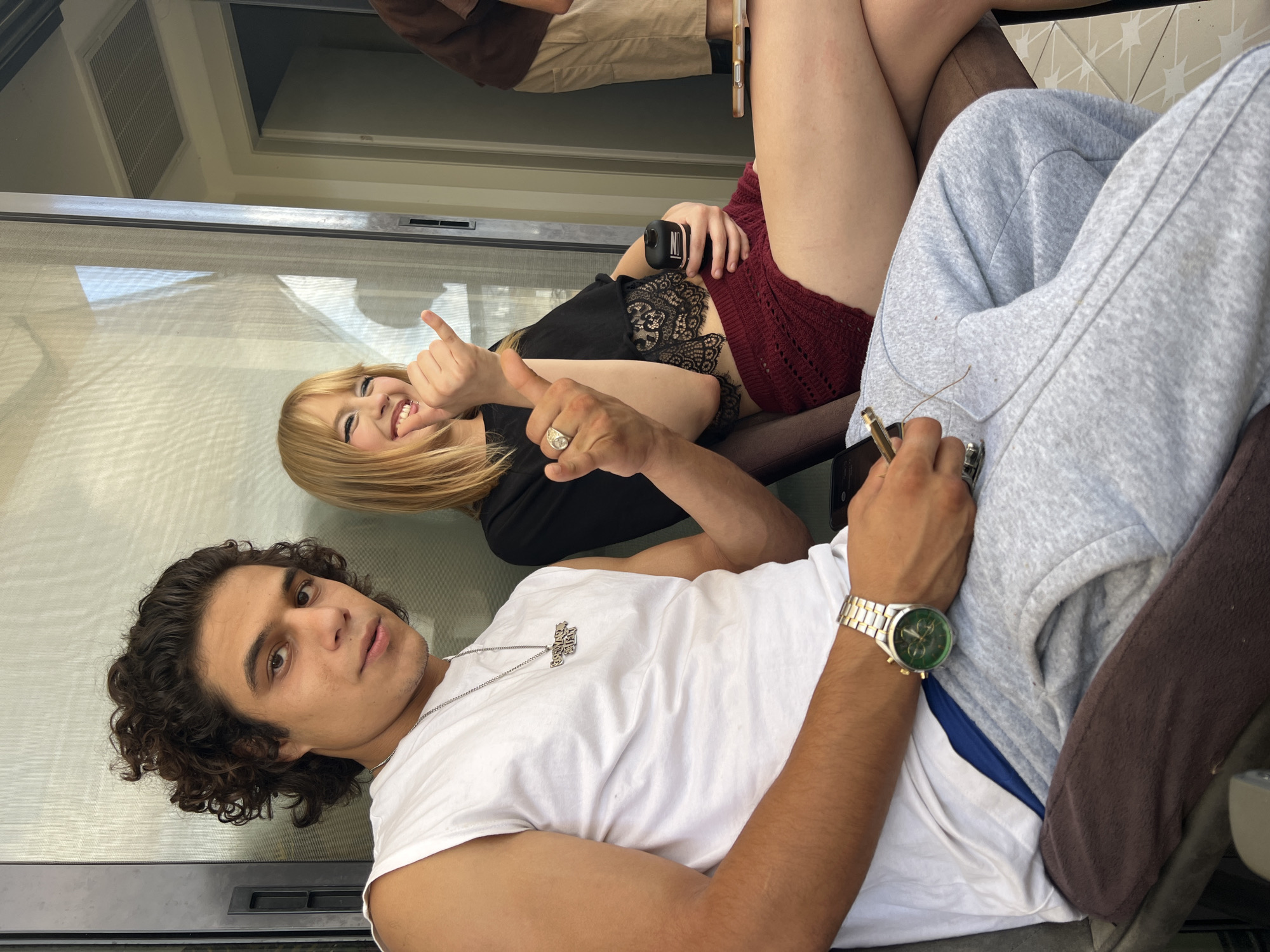 The media file shows two people, a man and a woman, in what appears to be an outdoor or semi-outdoor setting. The man is in the foreground, wearing a white t-shirt and a silver chain with a pendant. He has curly dark hair and is looking towards the camera with a relaxed expression. On his wrist, he wears a gold and silver watch with a green face. He is holding a cigarette or joint.
The woman is behind him, and partially visible. She has blonde hair and is wearing a black top with lace detailing and a red crocheted skirt. Her right hand is giving a thumbs-up. She is holding a black cylindrical object in her left hand. Both individuals appear to be casually dressed, suggesting a relaxed social gathering or downtime.
The background shows a window or glass door, with some reflections visible. The overall lighting suggests it is daytime, possibly late afternoon or early evening, given the warm tones. The setting appears to be a residential area, possibly a balcony or patio. The atmosphere seems informal and friendly, with the woman giving a thumbs-up indicating a positive mood. The location context provided is Kiryat Bialik, Israel. No other visible text is present in the image.