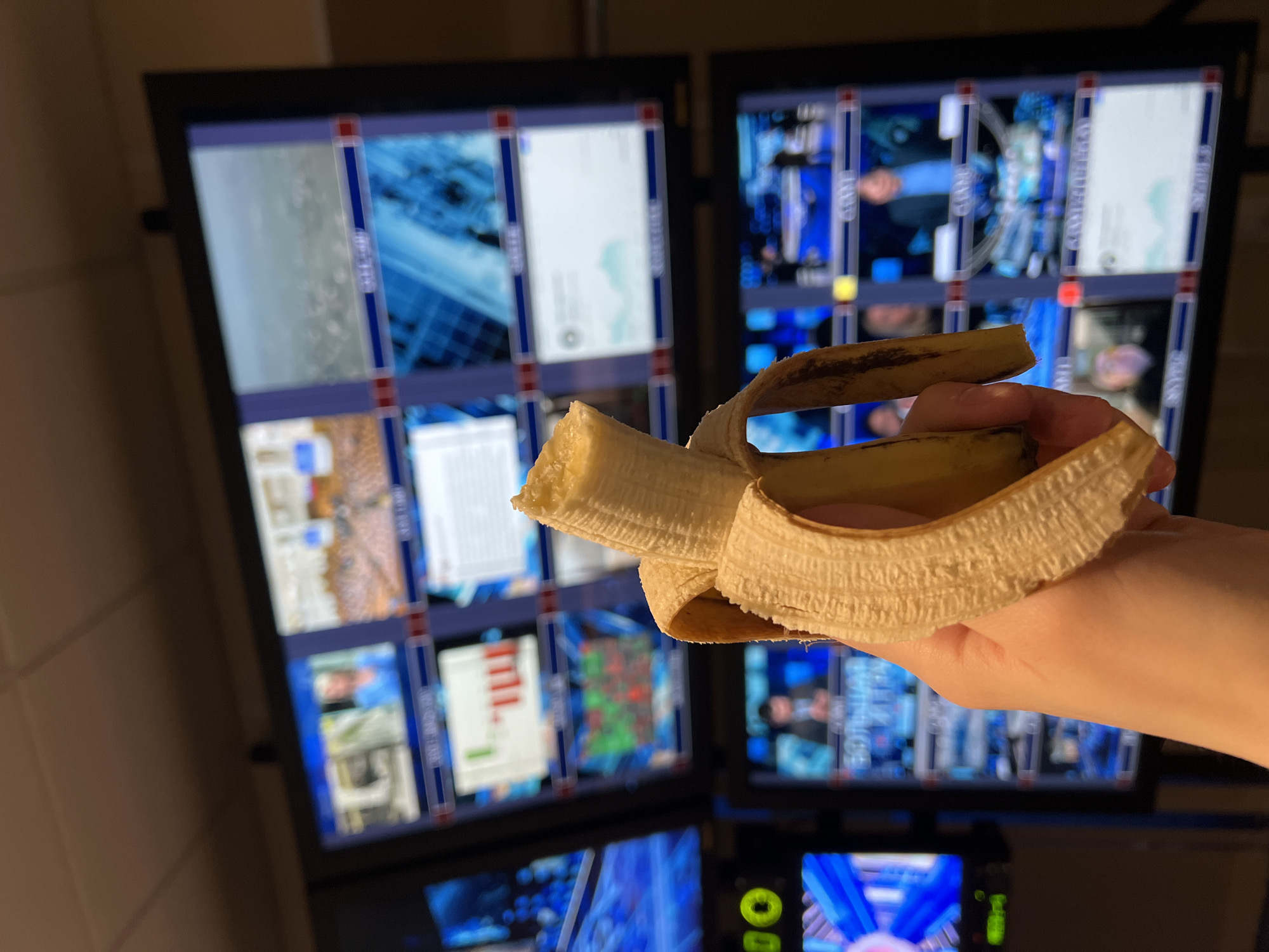 A peeled banana is being held in a hand in front of two large monitors displaying multiple video feeds. The video feeds appear to show news broadcasts, weather maps, and potentially surveillance footage. The location cannot be determined from the information given.