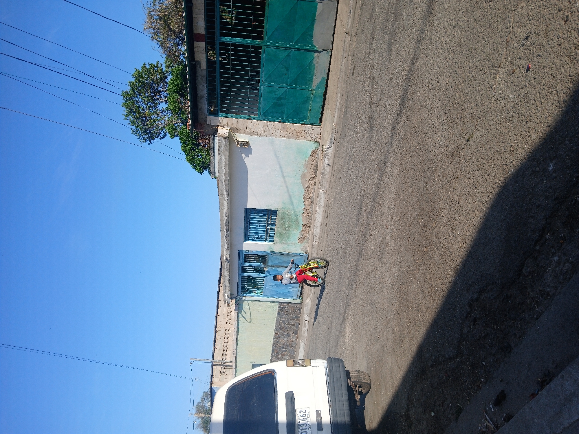 A street scene in Porlamar, Venezuela, features a boy on a yellow and black bicycle wearing a light grey shirt and red pants riding on the right side of a paved road. The road is bordered by a building on the left, with a white and light blue facade, a dark green metal gate and a small tree overhead. The sky is bright blue, with power lines visible in the upper left corner. The rear of a white van is visible in the lower left corner. The pavement is shadowed on the right side of the road.