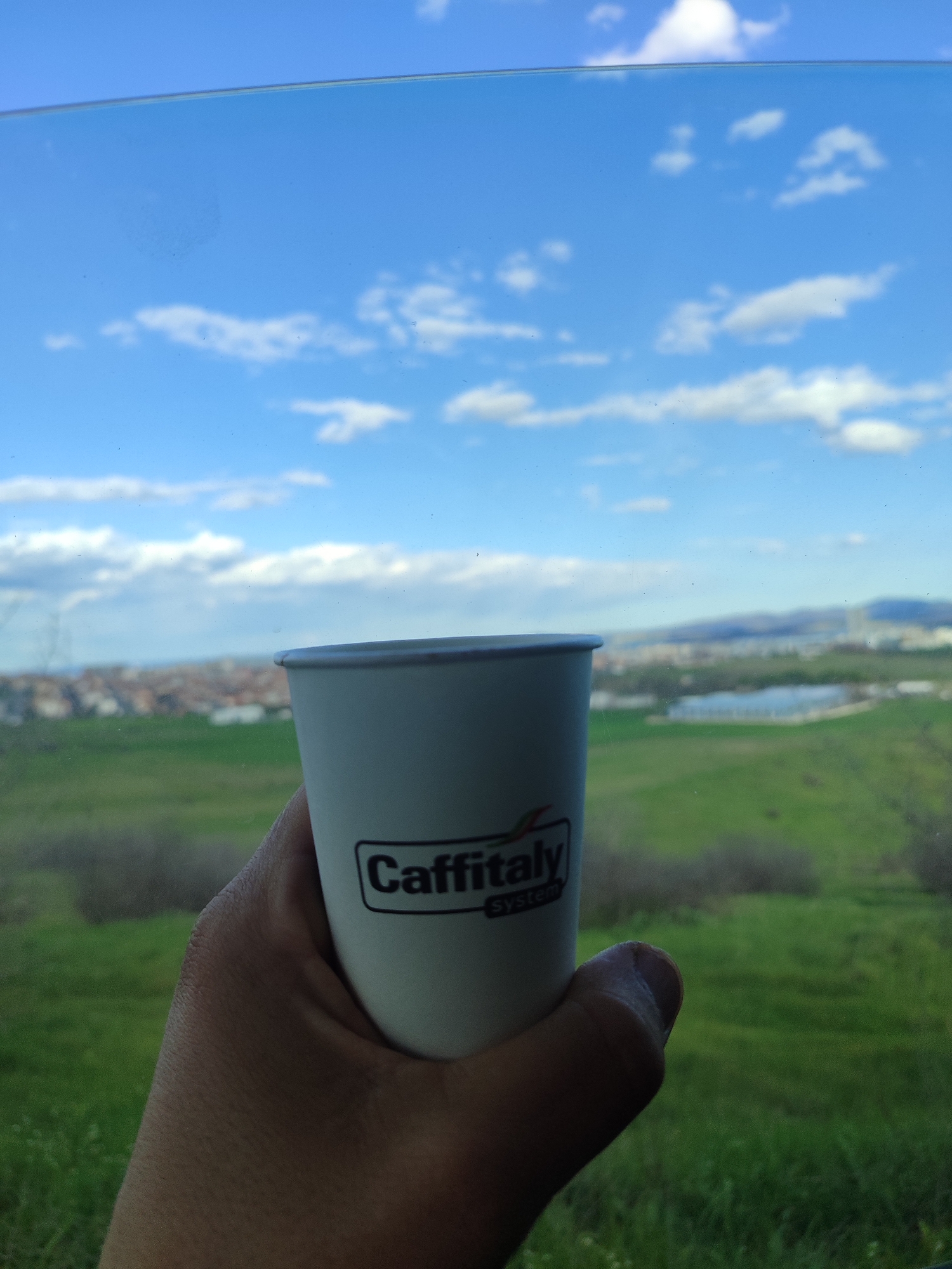 A person is holding a white paper cup with a "Caffitaly System" logo against a blurred background of a green field, buildings, and a blue sky with white clouds, visible through a window. The location is most likely in the city of Burgas, Bulgaria, based on the landscape features and architecture visible in the background.