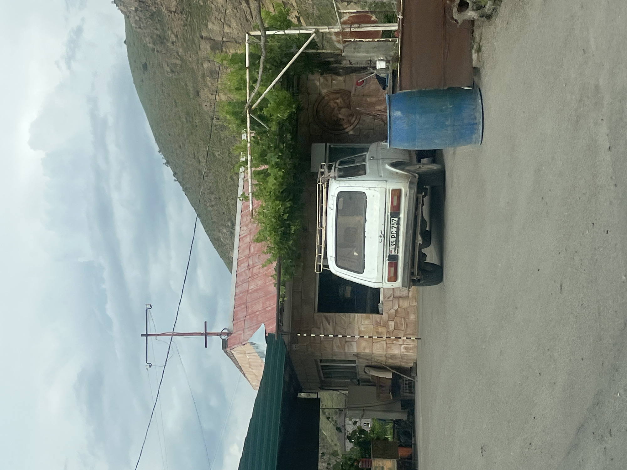 This image depicts a daytime scene in Chiva, Armenia, under a cloudy sky. The primary subject is a white Lada Niva SUV, equipped with a roof rack, parked on the right side of an asphalt road. The vehicle faces away from the viewer, and its rear license plate clearly displays "35 PC 752," consistent with Armenian registration.
Adjacent to the parked car, on the far right, a blue plastic barrel is visible. To the right of the road stands a building featuring a stone-faced exterior and a reddish-brown tiled roof. A metal framework, possibly a pergola or support structure, is erected in front of this building, partially covered by green foliage. Within the visible area behind this frame, a decorative circular relief or carving can be discerned on the building's wall, alongside what appears to be a red broom. A section of another building, featuring a dark green roof, is visible on the far left of the frame.
The background on the left is dominated by a large, steep, and verdant hillside or mountain, characterized by rocky outcrops under the overcast conditions. Against the sky on the far left, a utility pole with cross-arms and power lines is visible. The scene is static, showing no active events or people. The overall impression is of a quiet street in a mountainous or rural settlement.