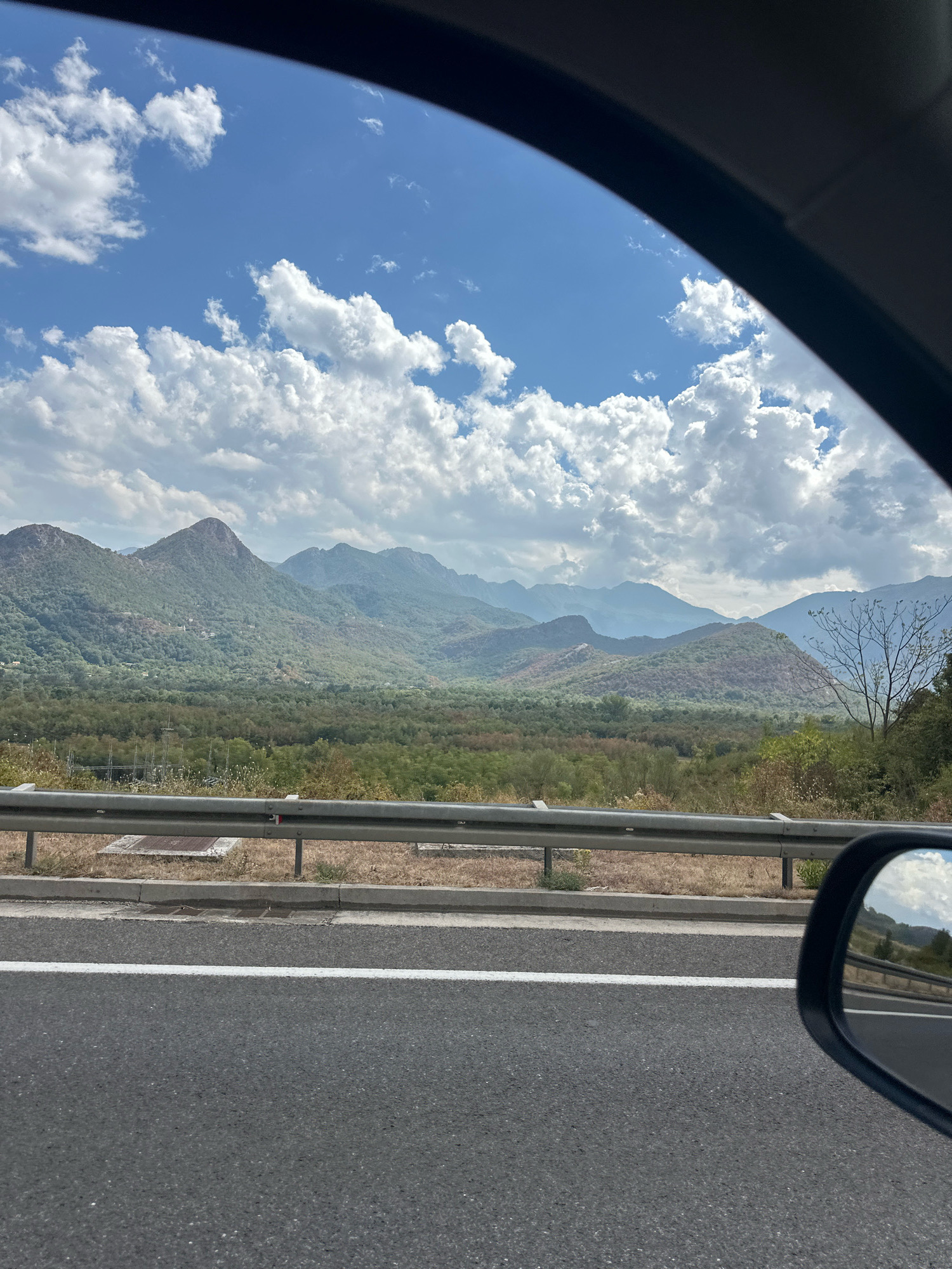 The image captures a scenic view from a car traveling along a highway, with a mountainous landscape and a blue sky filled with fluffy white clouds. The foreground features the asphalt road with a white lane marking and a metal guardrail. Beyond the guardrail, dry grass and some vegetation are visible. In the middle ground, a dense forest covers the rolling hills, leading up to the majestic, tree-covered mountains in the background. The mountains exhibit varying shades of green and brown, with some appearing more arid than others. The sky is a vibrant blue, dotted with scattered clouds, suggesting a clear and sunny day. The reflection in the car's side mirror shows a similar landscape, indicating the direction of travel. The scene appears to be located in a rural or natural area, likely near Sotonići, Montenegro, given the characteristic mountainous terrain.