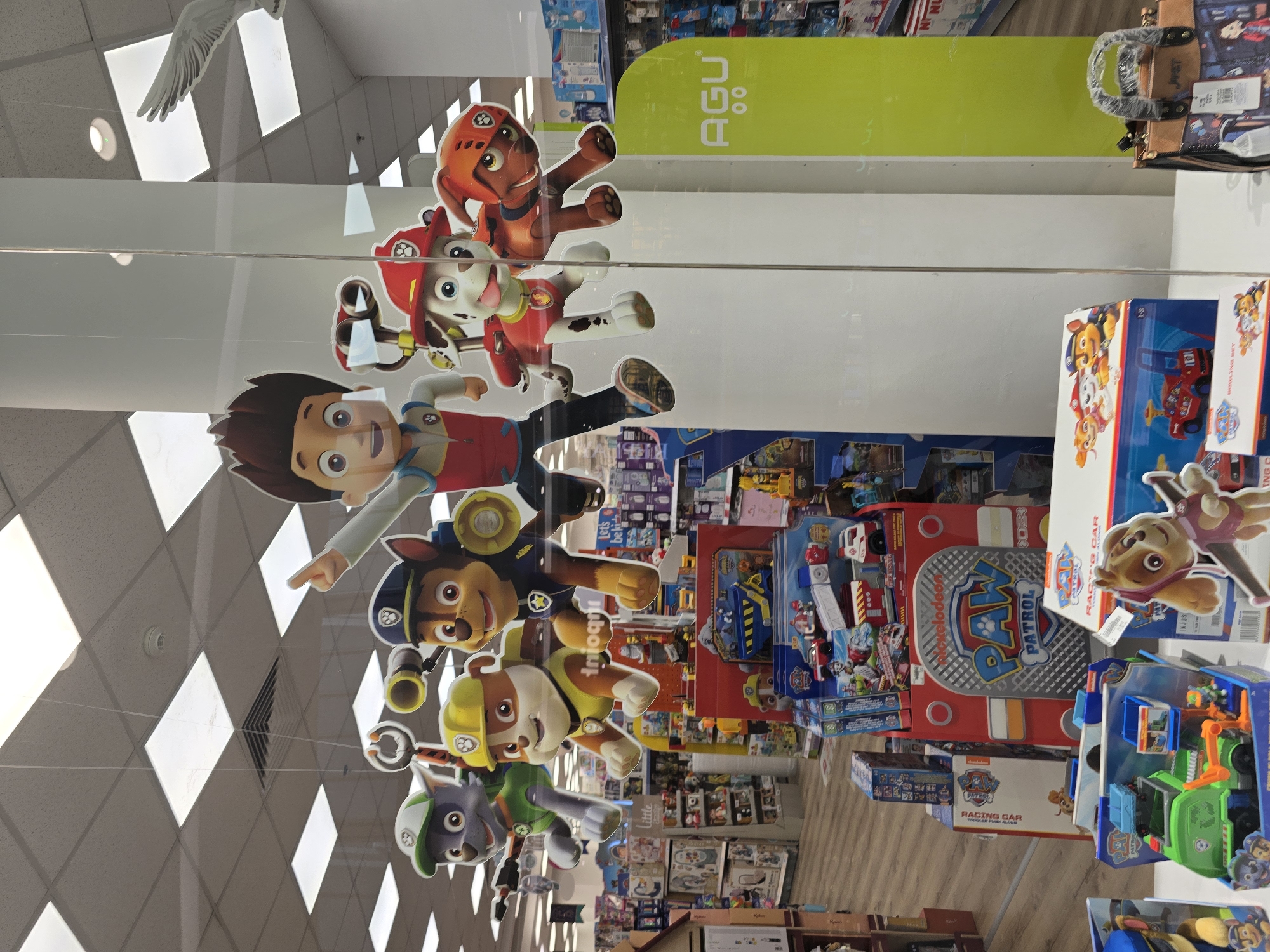 The image shows a store window display featuring characters from the Paw Patrol franchise. The characters, Chase, Marshall, Rubble, Skye, and Rocky, are depicted as cardboard cutouts hanging from the ceiling. In the foreground, several Paw Patrol toy sets are displayed on shelves, including a fire truck and a recycling truck. A purse with a Paw Patrol theme is also visible. The background reveals aisles of merchandise, typical of a toy store. The store is located in Plovdiv, Bulgaria, based on the text "Plovdiv" visible on a sign in the background. The lighting suggests it is daytime, and the scene is well-lit, likely from overhead fluorescent lights.