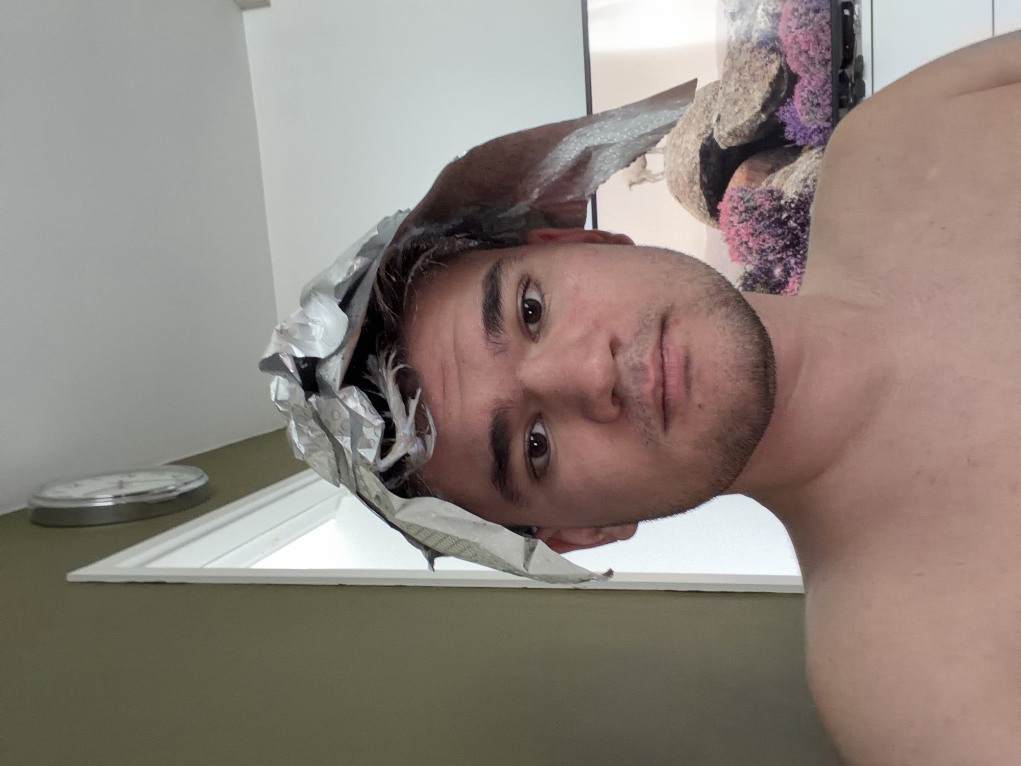 A medium shot photograph shows a man with short brown hair and a light beard, looking at the camera. He has aluminum foil wrapped on the top of his head, presumably for a hair treatment. The man appears shirtless. In the background, a window is visible with a clock on the wall to the left. To the right, a decorative coral-like object is visible. The location appears to be indoors, possibly a bathroom or living room. There is no information in the image to suggest the location is in Sofia, Bulgaria.