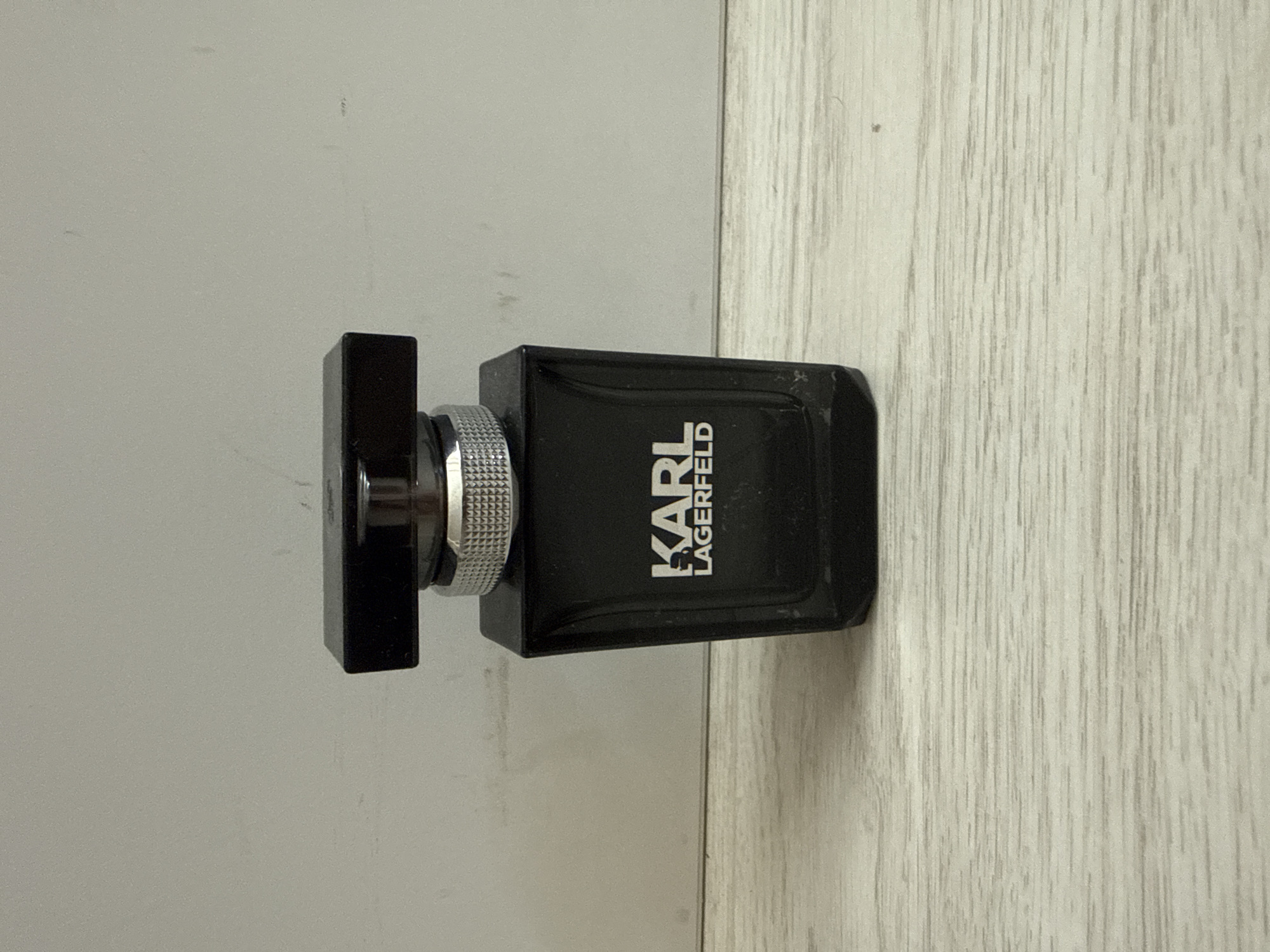 The image shows a black perfume bottle with the logo "KARL LAGERFELD" visible on its front. The bottle is made of glass and has a silver-colored cap with a textured grip. It is placed on a light-colored wooden surface, possibly a table or shelf. The background is a plain, light gray wall. There are no people or animals in the image, and no visible interactions are taking place. The scene appears to be indoors, with neutral lighting. The location is not specified in the image, and therefore cannot be determined to be in Burgas, Bulgaria.