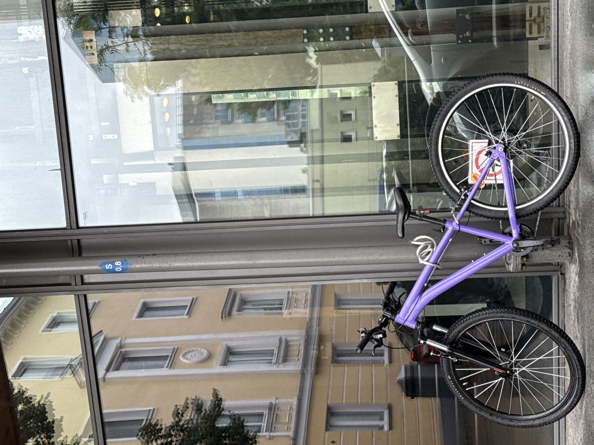 A purple bicycle is parked next to a modern building with large glass windows. The bicycle has a black frame, black tires, and silver spokes. It is secured with a silver U-lock. In the reflection of the glass windows, a yellow building with multiple windows is visible, as well as trees and other buildings. A small blue sign with white text that reads "S 0,8" is attached to a pole next to the bicycle. The ground is wet, suggesting recent rain.