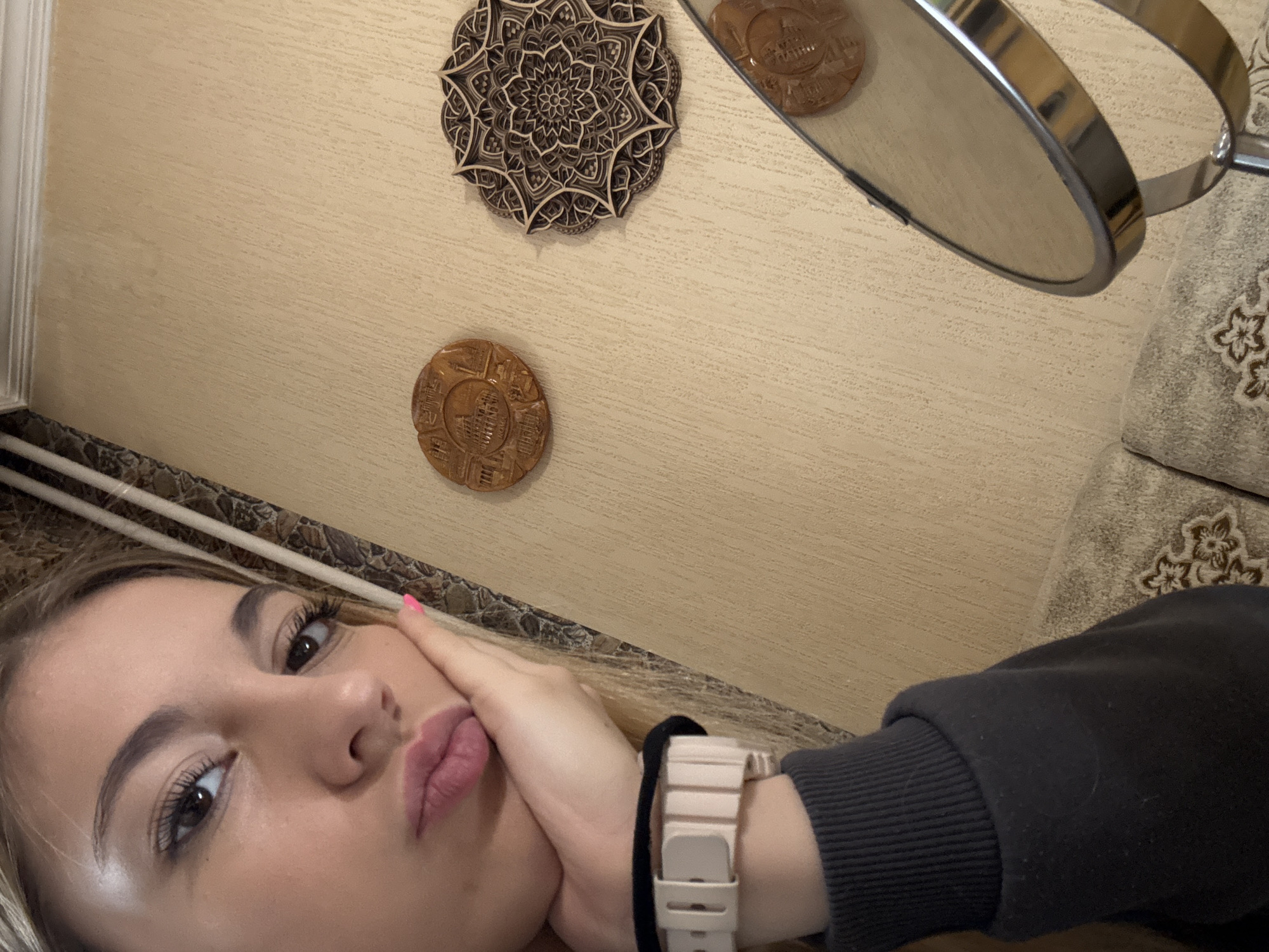 A close-up, tilted selfie shows a person with fair skin, long eyelashes, and pink lipstick, holding their face with their left hand, which is adorned with a pink-banded wristwatch and a black elastic band. They wear a dark gray sweatshirt. The background consists of a light-colored wall with wood-grain texture and three circular wall decorations: a mandala-style wooden carving, a round wooden plaque with an etched image, and the reflection of the same wooden plaque in a silver-rimmed mirror. A patterned couch is partially visible in the upper right corner. Based on visual evidence, the location of the image cannot be determined, and thus, a precise location within Sofia, Bulgaria cannot be provided.
