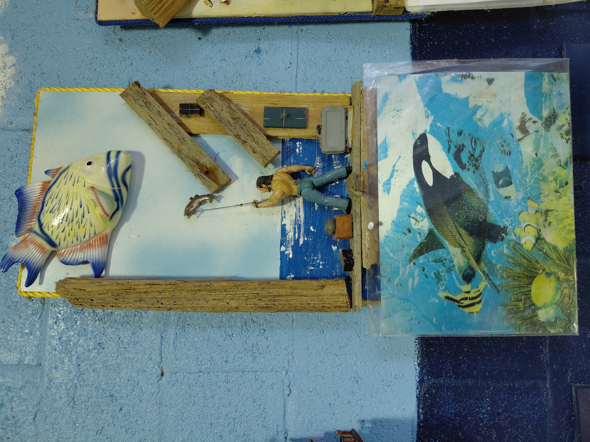 The image shows a diorama or art piece. The scene depicts a fisherman in a small, stylized wooden boat pulling in a fish, with a large ceramic fish figure positioned to the side. The boat and surrounding "water" are constructed from wood, and a small structure resembling a shack or cabin is also included. Next to the boat scene is a framed image of an orca and various other fish. The entire composition is set against a light blue wall, which has a slight texture suggesting it may be concrete or plaster. The lighting is somewhat even, with no strong shadows to suggest a single prominent light source. The location is indicated as Cutler Bay, United States of America.