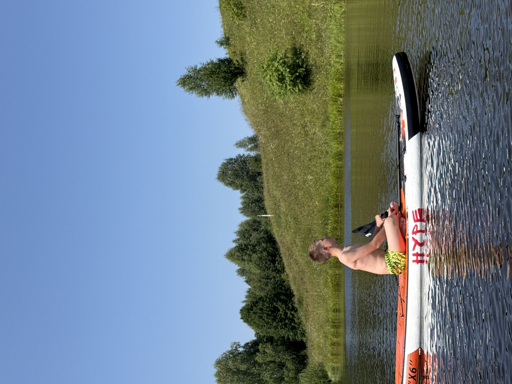 A young man is sitting on a paddleboard in a body of water. The paddleboard is white with orange and black accents, and text that reads "HYFE" is visible. He is wearing yellow patterned swim trunks and appears to be looking up and to his right. The water is dark and rippled, suggesting a calm day.
Behind the paddleboard, a grassy hill slopes upwards and is dotted with trees. The sky is clear and blue, indicating fair weather. The scene appears to be set in a natural, outdoor environment, possibly a lake or slow-moving river. The lighting suggests it is daytime, likely mid-morning to late afternoon. The overall mood is peaceful and recreational.