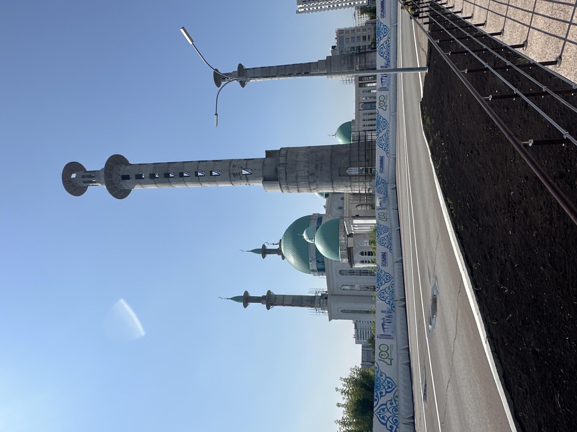 This is a daytime, eye-level, exterior shot of the construction of a mosque in Naberezhnye Chelny, Russia. The main subjects are the mosque's partially constructed minarets and domes, which are made of concrete and are topped with green domes and crescent moons. The largest minaret, on the right, is significantly taller than the others and features multiple levels with open windows. The mosque itself has white walls and arched windows, with prominent green domes. In front of the mosque is a wide, paved road with white lane markings and some puddles on its surface. A low concrete barrier separates the road from a sidewalk area. This barrier is decorated with a repeating pattern of blue ethnic motifs and the logos of "400" and "KAMAZ." The background includes trees and other buildings, suggesting an urban environment. The sky is clear and blue with a faint, lens-flare-like reflection. There are no people visible in the frame.