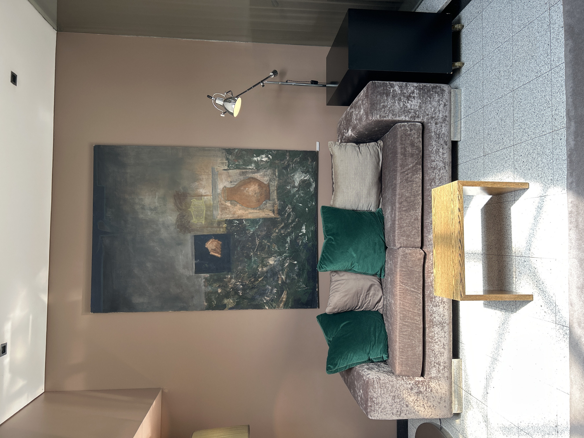 A light-filled, modern living room features a plush, taupe-colored sofa with velvet upholstery. Two emerald green velvet throw pillows and two taupe linen throw pillows are arranged on the sofa. In front of the sofa sits a small, light wood coffee table. A large, abstract painting with a dark, textured background and muted tones hangs on the taupe wall behind the sofa. To the right of the painting, a sleek, black cabinet stands against the wall, topped with an adjustable, silver metal floor lamp. The floor is a speckled terrazzo tile.