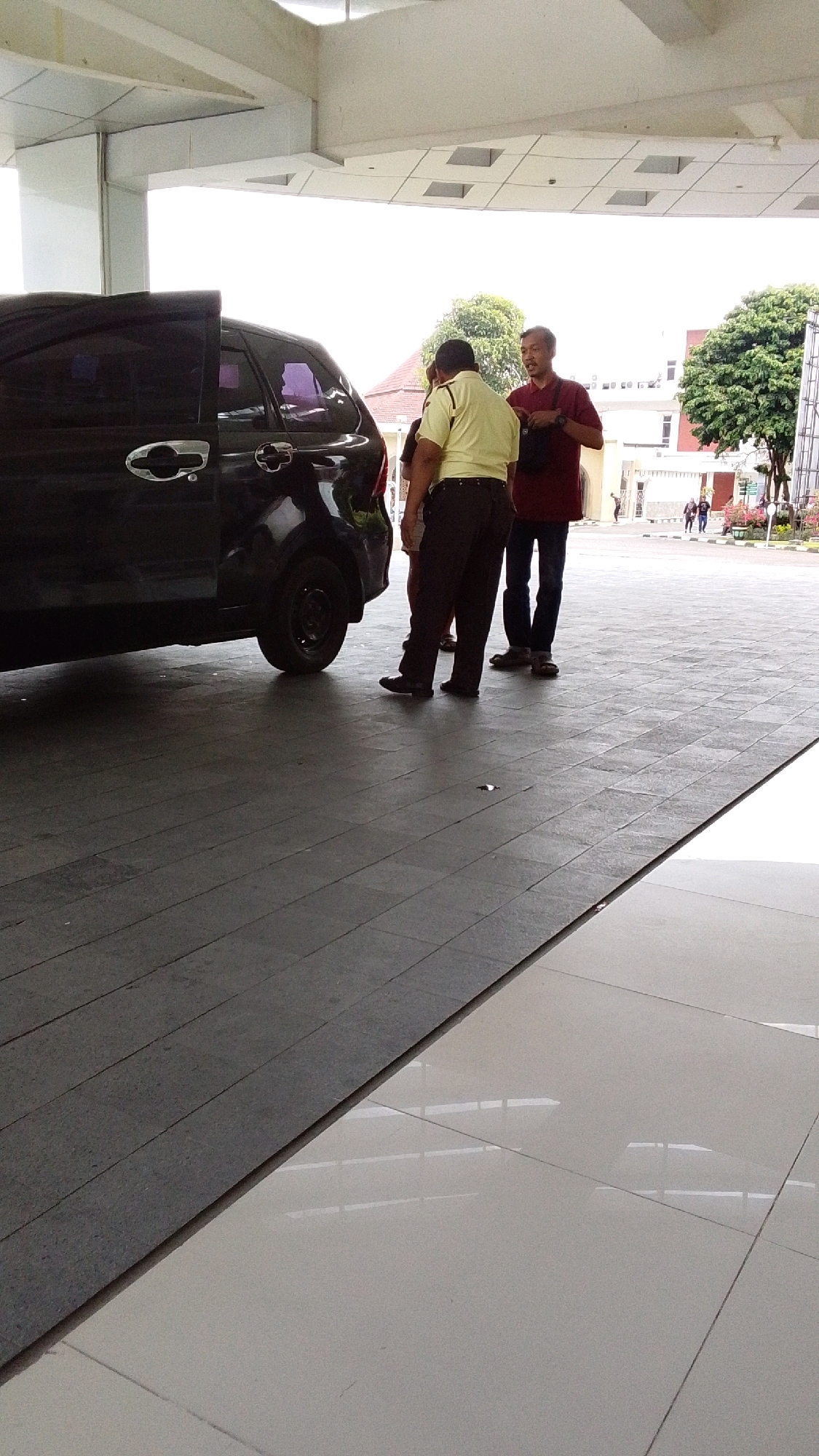 Two men are standing next to a black car parked under a white overhang in what appears to be an outdoor public space. One man, on the left, is wearing a light yellow short-sleeved shirt and dark pants. He is facing away from the camera and appears to be looking at the car. The other man, on the right, is wearing a dark red long-sleeved shirt and dark pants. He is facing the first man and appears to be in conversation. The car has its rear passenger door open. In the background, there are buildings and trees. The ground is paved with large, dark gray square tiles. The area is well-lit by natural light, suggesting it is daytime. This scene could be located at a university campus or a public facility in Pabelan, Indonesia, given the architectural style of the buildings and the tropical foliage.
