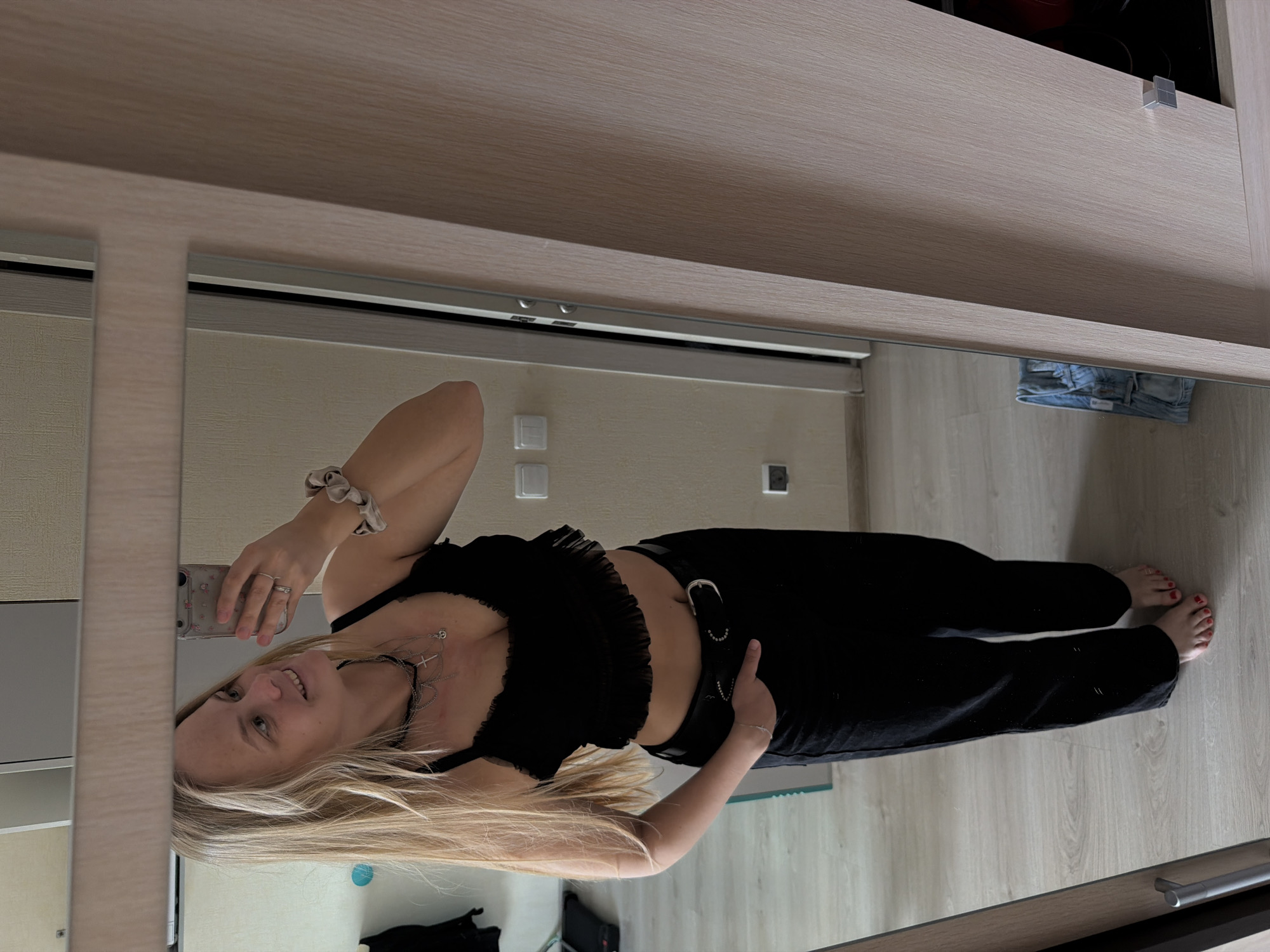 A full-body mirror selfie shows a young woman posing indoors, rotated 90 degrees counter-clockwise. The woman, with long blonde hair, a fair complexion, and a slight smile, is the central subject. She is wearing a black, ruffled crop top that exposes her midriff, black pants, and a black belt with a prominent silver buckle and metallic embellishments. She has multiple silver necklaces, including one with a cross pendant, and a ring on her left hand. A light-colored scrunchie is on her left wrist, and she holds a smartphone with a patterned case in her left hand to capture the photo. Her right hand is casually placed on her belt, near her hip. Her bare feet are visible, with red-painted toenails, resting on a light wooden floor.
The reflected setting appears to be a modern room, likely a residence in Izhevsk, Russia, with light wooden flooring and plain, light-colored walls. In the background reflection, white light switches are visible on the wall. Further back on the floor, a pair of folded light blue jeans is seen, along with a dark blue object, possibly a bag. The top edge of the mirror is framed by light wooden cabinetry or shelving. The room is well-lit, suggesting either daytime or bright artificial lighting. The woman appears confident and content, striking a casual pose for the mirror selfie. No legible text is visible in the image.
