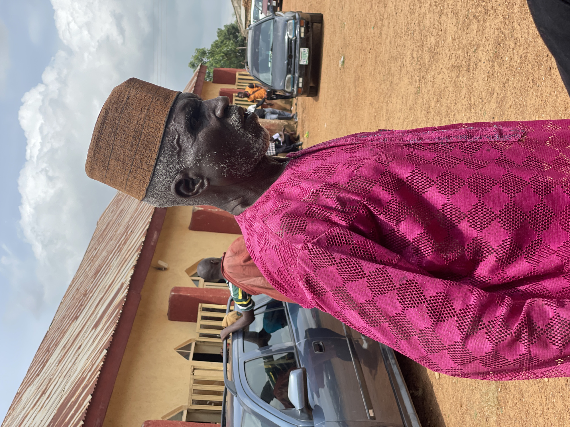 This is a vertical, medium shot of an elderly man in profile, facing left. He wears a brown, textured cap and a vibrant magenta, lace-like fabric garment. His weathered face, with a white beard and mustache, is turned upwards, and he appears to be looking at something in the sky. The background suggests an outdoor gathering in Ban, Nigeria.
Behind the man, a single-story building with a corrugated metal roof and yellow walls is visible. Several people are gathered around a dark gray car, which is parked on a dirt road. One person in an orange shirt is leaning on the car. The ground is covered in reddish-brown dirt. The sky above is partly cloudy with bright white clouds against a pale blue sky. The scene appears to be set during the daytime, possibly late morning or early afternoon, given the brightness of the light. The overall mood is relaxed and informal, perhaps a community event or a social gathering. There is no visible text in the image.