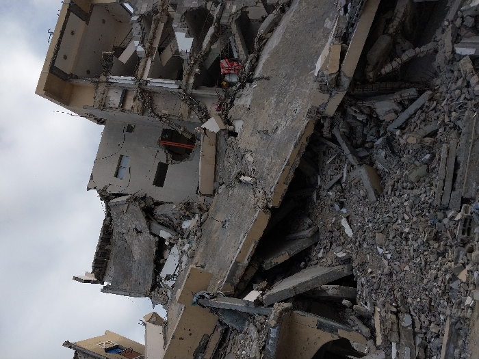 A partially collapsed building, likely due to damage from a catastrophic event. Debris, including concrete slabs, rubble, and twisted metal, is scattered across the foreground. The remaining structure reveals fragmented rooms and open spaces, showcasing the building's ravaged interior. The sky is overcast, adding to the somber atmosphere.
