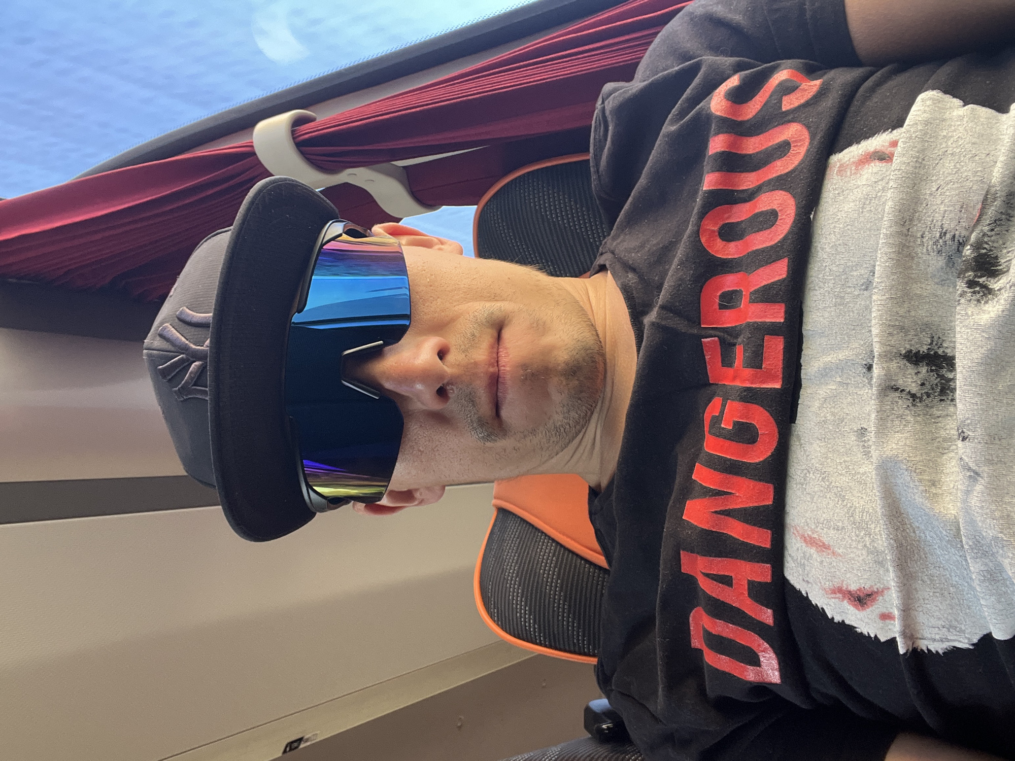 A person is sitting in a chair on what appears to be a train or bus in Chaikovsky, Russia. The individual is wearing a black baseball cap with a symbol on the front, mirrored sunglasses, and a black t-shirt with the word "DANGEROUS" printed vertically in red letters along with a white graphic. Red window shades are visible above the person, and a seat with orange accents is behind them. The window shows a view of the sky, and the overall setting suggests a journey or commute.