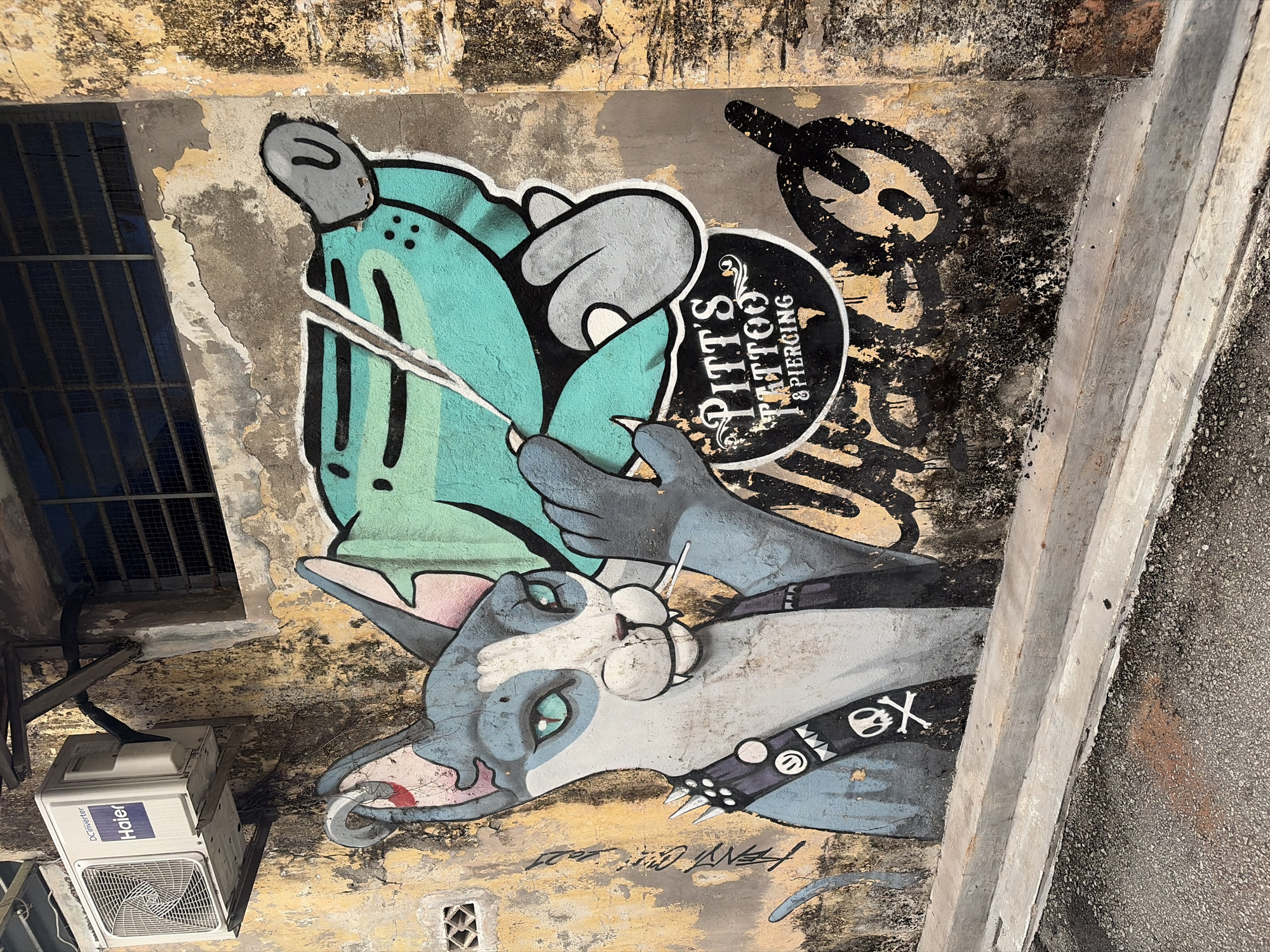 The image depicts a section of an outdoor wall in an urban environment, likely a street. The wall itself is weathered, with patches of yellow and grey paint, and areas of cracked and peeling plaster, suggesting age and exposure to the elements. The lower portion of the wall is bordered by a concrete curb and asphalt road.
Dominating the wall is a colorful graffiti mural. The central subject is a stylized cat character rendered in shades of grey, blue, and white, with striking green eyes. The cat is depicted wearing a dark collar adorned with spiked studs and a patch featuring a smiling face emoji and a skull and crossbones symbol. The cat is holding a large, cartoonish green object, possibly a piece of food or a toy, in its paw, with a cigarette or similar object in its mouth.
To the right of the cat mural, there is a black circular logo with white text that reads "Pitt's Tattoo & Piercing". Above the cat mural, an air conditioning unit, branded "Haier", is mounted on the wall, supported by metal brackets. The AC unit appears functional. To the right of the AC unit, a barred window is visible. The overall impression is a piece of street art on a distressed urban wall, adjacent to commercial signage and some basic urban infrastructure. The specific location is unknown.