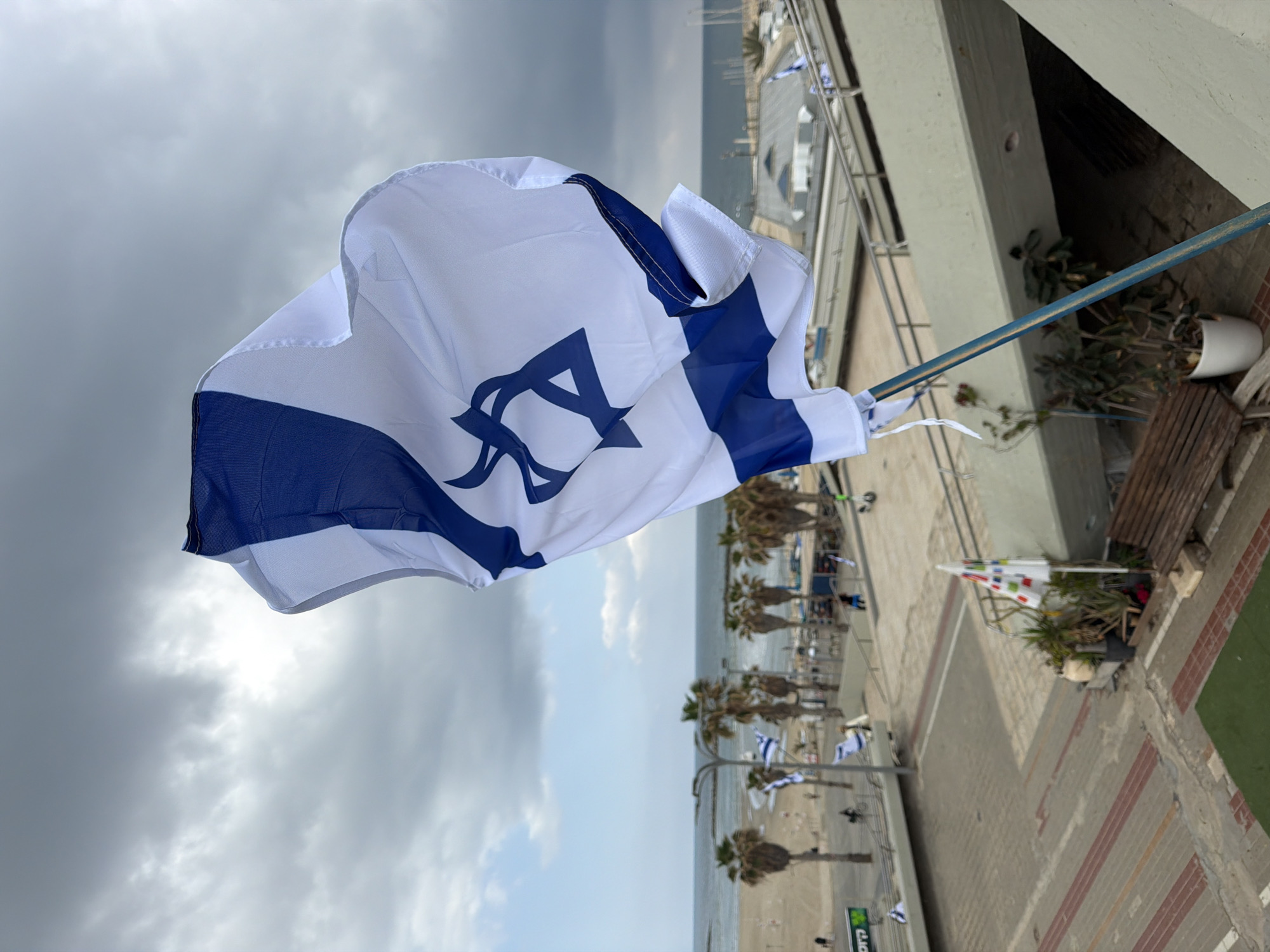 The image shows an Israeli flag waving prominently in the foreground, its blue Star of David and stripes visible against a white background. The flag is attached to a pole extending from a balcony.
In the background, there's a view of what appears to be the Tel Aviv beach promenade. The beach itself, the sea, and a cloudy sky are all visible. A row of palm trees lines the promenade, and smaller Israeli flags are seen on street lamps or poles along the walkway. A few people can be spotted walking along the promenade.
The balcony from which the image is taken is part of a building with a concrete structure. There is a bench, some plants, and other decorative items on the balcony.
