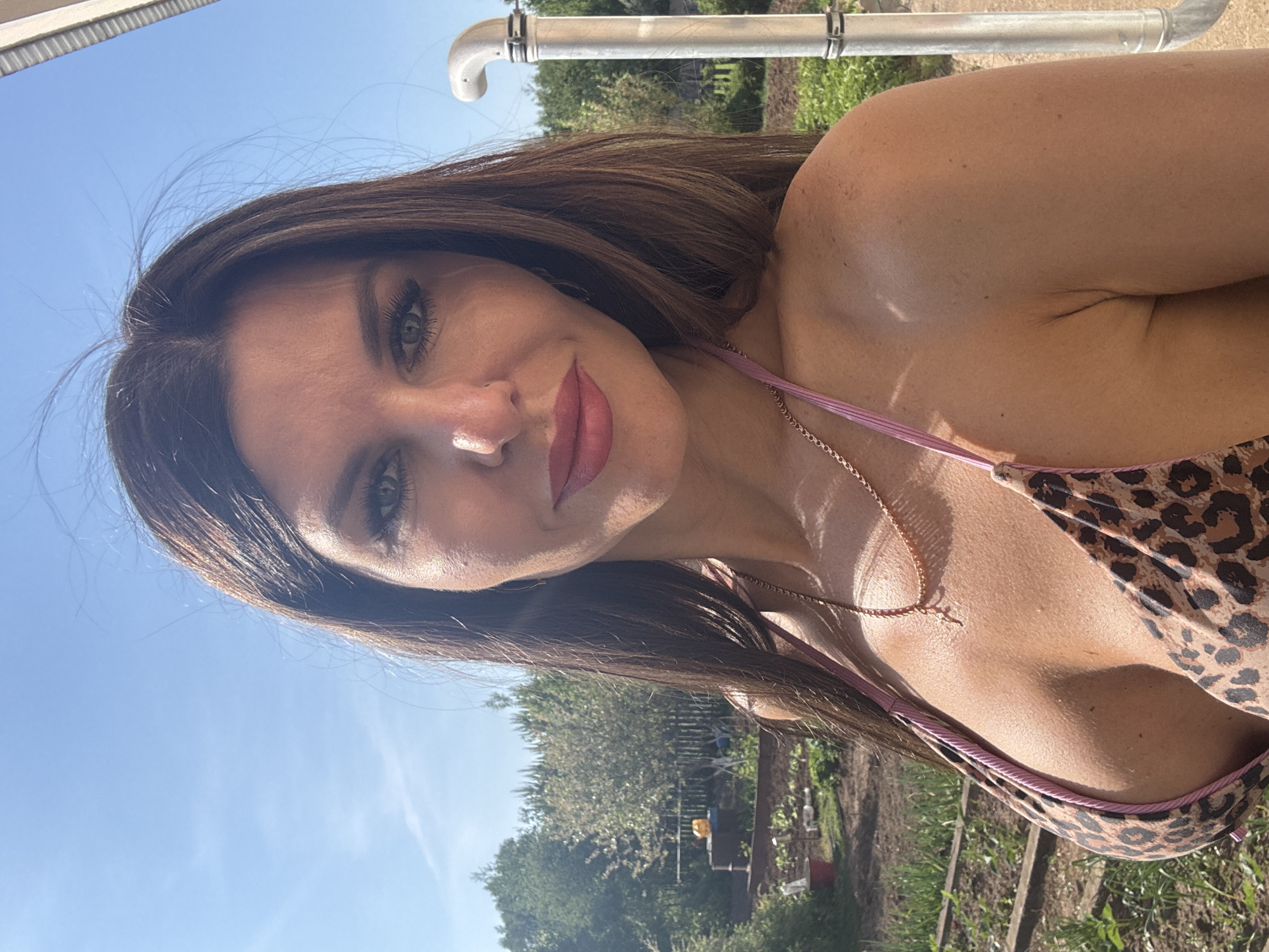 A woman is shown in a selfie, wearing a leopard-print bikini top with pink straps and a delicate gold necklace. She is outdoors, with a blue sky visible in the upper left. Behind her are trees, green vegetation, and a fence, suggesting a garden or park setting. A silver pipe structure is visible above her head. Based on this information, the image does not provide sufficient detail to confirm the location in the city of Dubovaya, Russia.