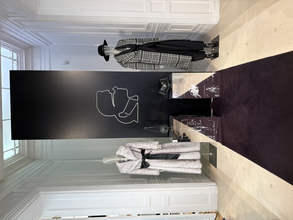 This is an interior view of a fashion display within an ornate retail or exhibition space, likely in Paris, France, given the classical architectural details. The scene features white paneled walls with detailed moldings, a light-colored polished stone floor, and a translucent ceiling panel with decorative elements providing overhead light.
A tall, matte black rectangular panel stands centrally, adorned with a white linear outline depicting the profile of Karl Lagerfeld wearing glasses. A dark burgundy velvet runner extends from the foreground towards this central display. Flanking the black panel are two mirrored pedestals with dark purple sides featuring white line-art fashion sketches and "Karl Lagerfeld" branding. On top of the left pedestal rests a spherical, textured silver-toned handbag, while a black, geometric or faceted handbag sits atop the right pedestal.
Two transparent-headed mannequins are positioned on either side of the central display. The mannequin on the left wears a full-length, light grey faux fur coat with a wide collar, cinched at the waist with a black satin ribbon belt. The mannequin on the right is dressed in a black wide-brimmed hat, a long black and white plaid trench coat, black wide-leg trousers, and black ankle boots. White paneled doors are visible on both the left and right sides of the scene.