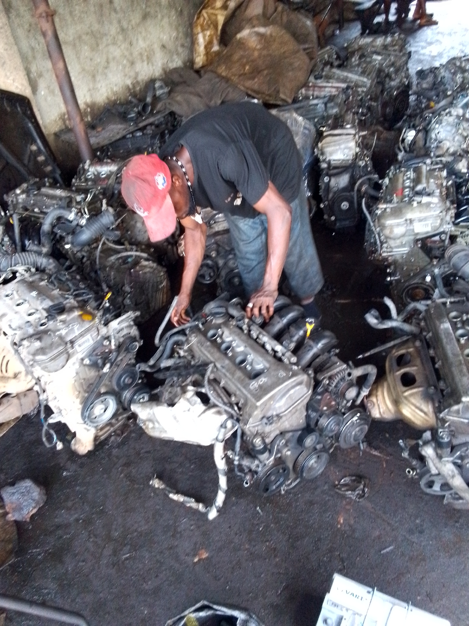 The image captures a man working amidst a dense collection of used car engines in what appears to be an auto spare parts yard or workshop, consistent with settings found in Onitsha, Nigeria. The man, wearing a dark t-shirt, blue trousers, and a red baseball cap, is leaning over one of the engines, seemingly inspecting or manipulating its components with his hands.
Numerous car engines, ranging in size and type, are scattered across the dark, oil-stained concrete floor. Many engines have visible accessories like timing belts, pulleys, and alternators, indicating they are either salvaged or awaiting repair/disassembly. The background features a rusty metal pole and bundles or bags, possibly containing other parts or materials, against a concrete wall.
The activity suggests the man is a mechanic or a vendor dealing with used engine parts. The lighting indicates it is daytime, likely an indoor or semi-covered space. The overall scene is cluttered and utilitarian, reflecting the nature of a busy auto parts environment. A white box with the partially visible text "VART" is situated in the bottom right corner of the frame. The man appears focused on his task.