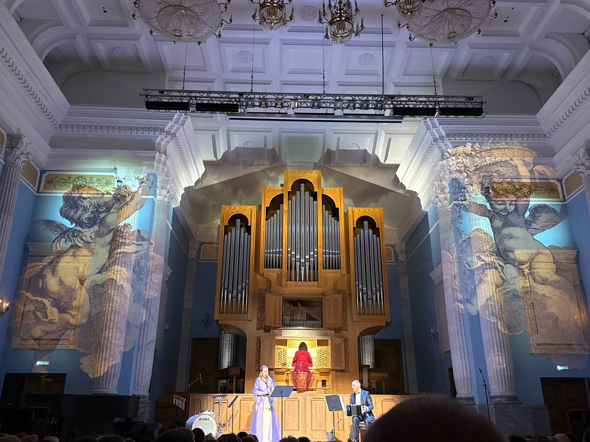 A concert hall stage is depicted, featuring a large organ as the central element. Two individuals are performing on the stage, with a third person seated at the organ console. The hall's architecture is ornate, with high ceilings, decorative molding, and large chandeliers. The walls are painted a light blue and adorned with projected images of classical artwork, possibly cherubs.
The performer on the left is a woman in a long, flowing lavender gown, playing a wind instrument. To her right, a man in a suit and bowtie is playing an accordion. Seated behind them, at the organ console, is a person in a red outfit, presumably the organist. In front of the performers, a drum kit is visible.
The audience is partially visible in the foreground, suggesting a live performance. The lighting on stage is bright, illuminating the performers and the organ. The overall atmosphere suggests a formal classical music event.
The image is taken from the audience's perspective, looking towards the stage. The venue is the Chelyabinsk State Academic Opera and Ballet Theatre in Chelyabinsk, Russia, as evidenced by the architectural style and the prominence of the organ, which is a significant feature of the theatre.