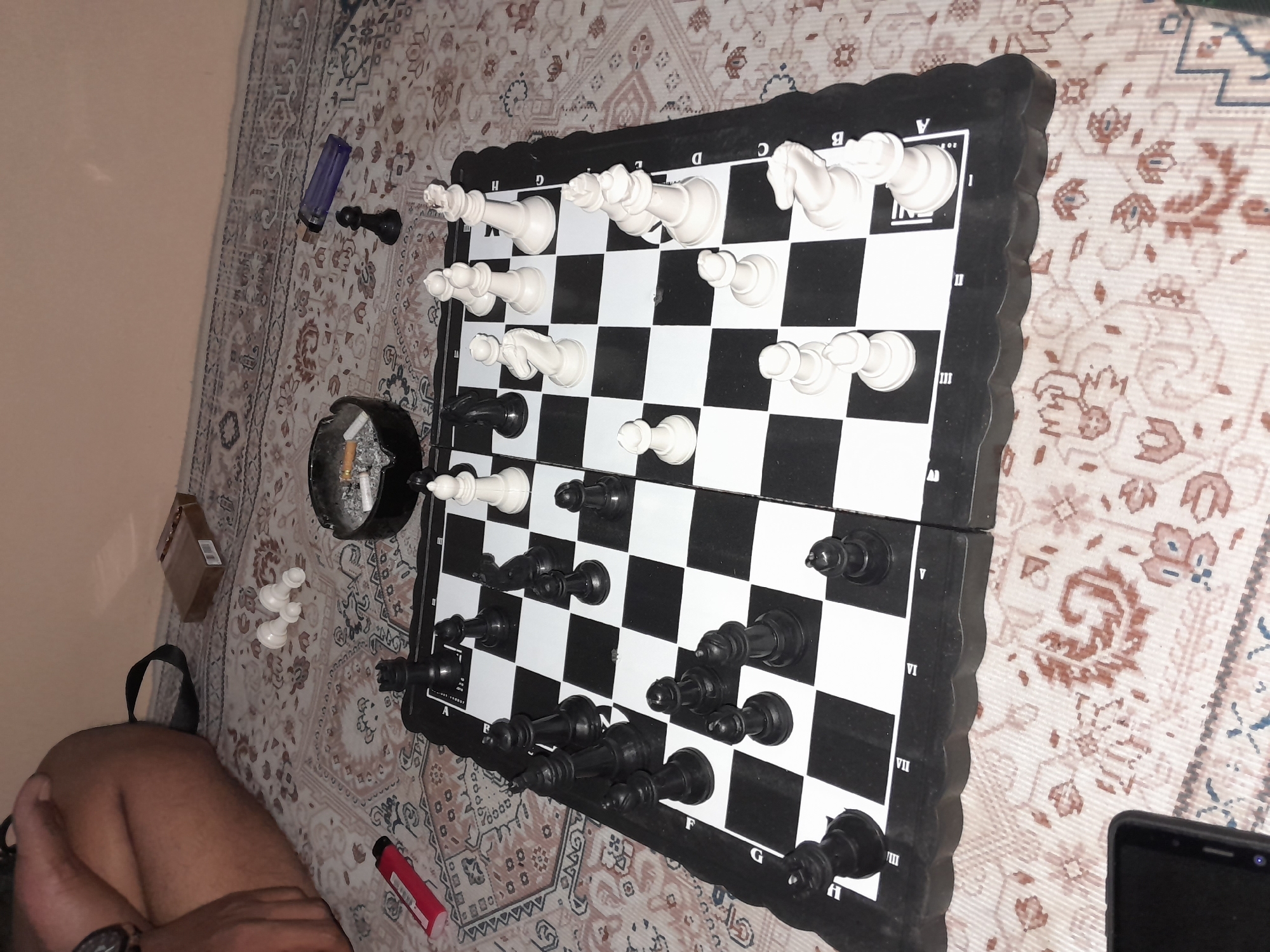 The image shows a chess board set up on a patterned rug. Black and white chess pieces are arranged on the board, indicating a game in progress. Scattered around the chess board are several objects, including a cigarette pack, an ashtray with cigarette butts, a red lighter, and a blue lighter. A person's legs are visible in the upper left corner of the image. The overall environment suggests an indoor setting, possibly a living room or a casual area. The image does not contain any specific visual cues that would place it in Tegal, Indonesia.