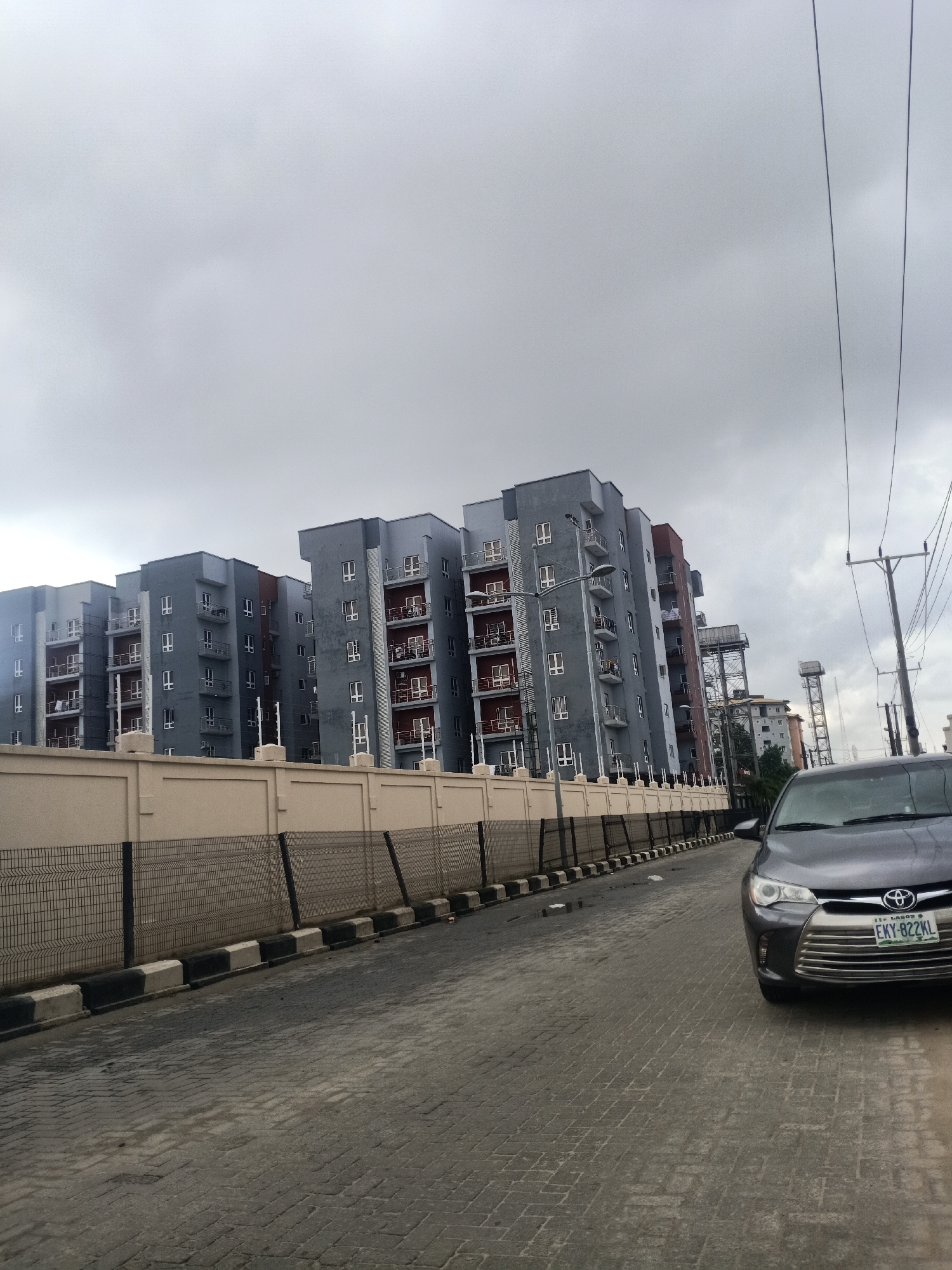 The image shows a street scene in Igboefon, Nigeria. Gray apartment buildings line the left side of the street, behind a beige wall topped with a black wire fence. The street is paved with interlocking pavers and edged with black and white painted curbs. A dark gray Toyota sedan is parked on the right side of the street, partially blocking the view. Utility poles and wires are visible against an overcast sky. In the distance, behind the apartment buildings, are water towers. There are no visible people in the image.