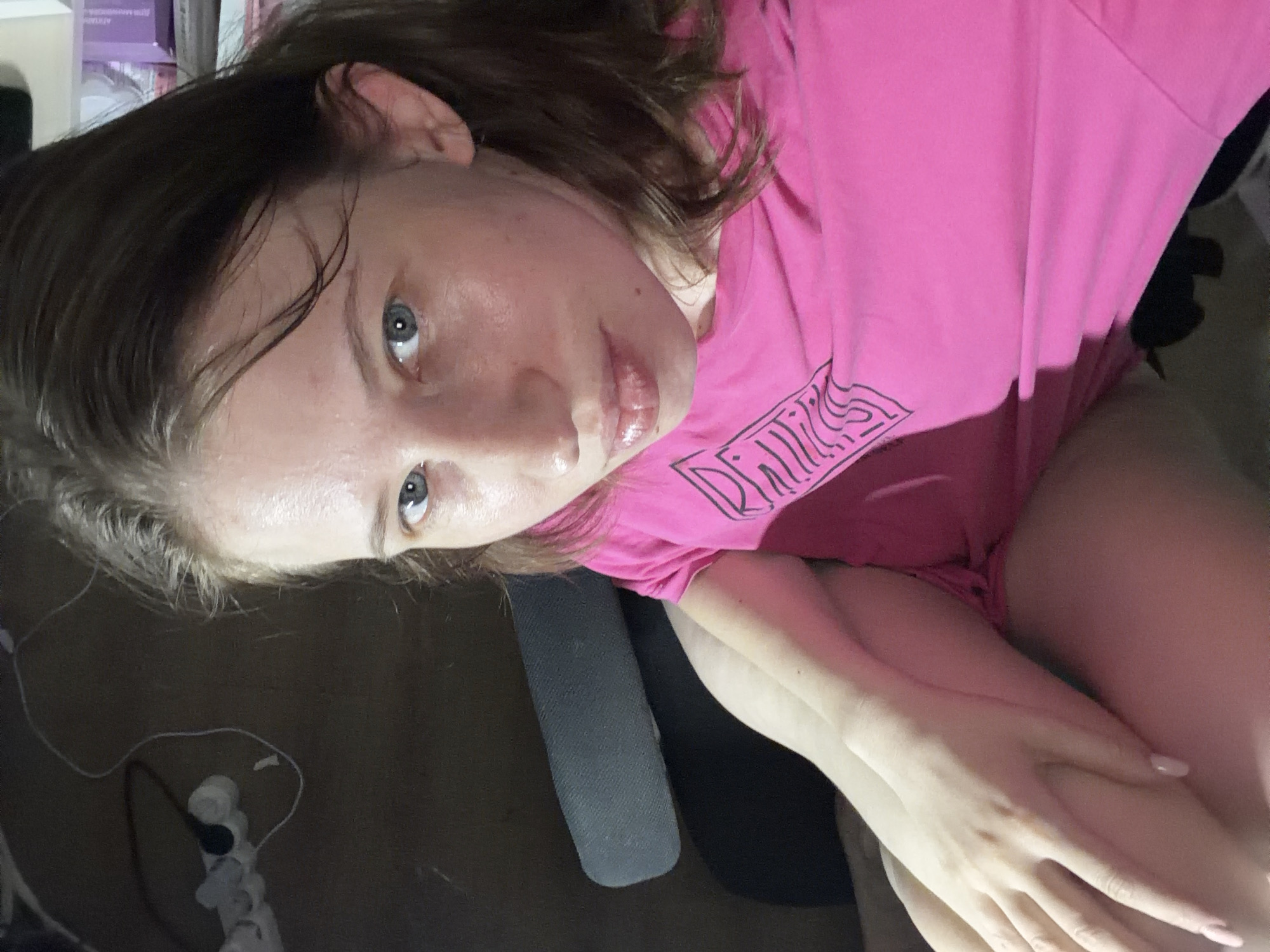 A young woman with blue eyes and light brown hair is captured in a close-up shot, looking directly at the camera. She is wearing a bright pink t-shirt with a black graphic design on it. Her arms are crossed, with one hand resting on her leg. The background is dimly lit and appears to be indoors, with some electrical cords and what looks like a desk chair visible. The image was taken in Dongducheon-si, South Korea.