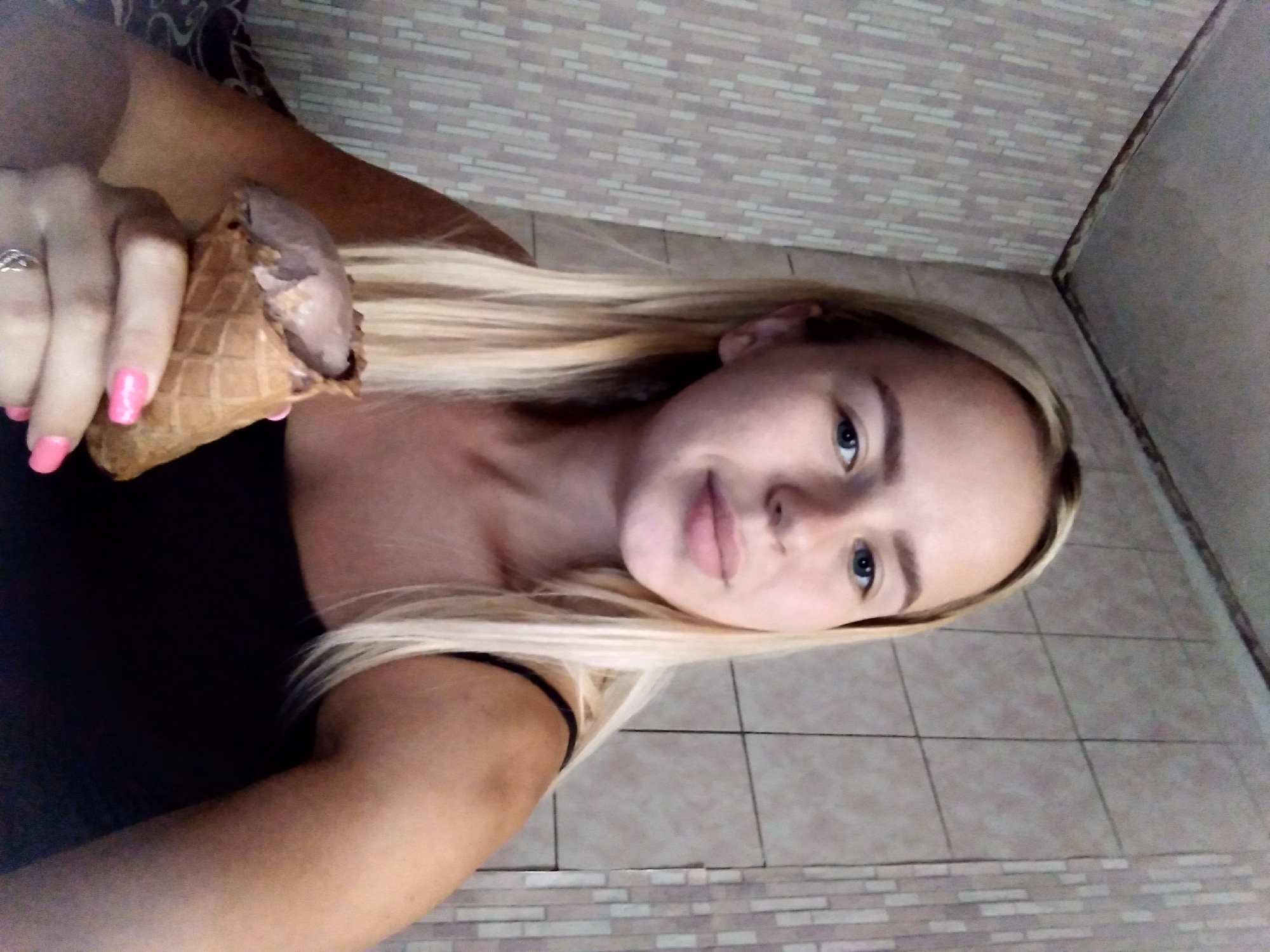 A young woman with blonde hair and blue eyes is sitting indoors and holding an ice cream cone. She is wearing a black tank top and a ring on her left hand. The background consists of tiled walls. The location is Radisne, Ukraine.