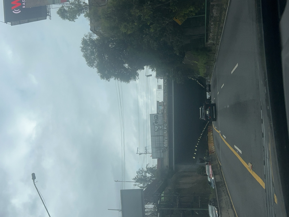 The image displays a multi-lane highway approaching a tunnel entrance under an overcast sky, captured from inside a vehicle. The road features a solid yellow line on the left and dashed white lane markers. A dark-colored SUV occupies the center lane, proceeding towards the tunnel. To the immediate left, a concrete barrier separates the main road from a shoulder or side lane where several vehicles, including a red car, are stationary. Yellow plastic crash barriers are positioned along the left edge of the active lanes.
An overhead concrete bridge, heavily covered in graffiti, spans the highway directly before the tunnel entrance. Above and to the left, an elevated metal structure, possibly a pedestrian walkway with stairs, is visible. Utility poles and multiple power lines crisscross the upper-mid section of the frame. The tunnel entrance itself is dark, with visible internal lighting. To the right of the tunnel, a steep, densely vegetated embankment rises, featuring numerous trees and foliage. A large billboard displaying "W RADIO" is partially visible high on the right, obscured by trees. Another billboard structure is partially seen on the far upper left. The sky is uniformly gray and overcast. The road surface appears damp. The scene is located on a highway in Unknown, Unknown.