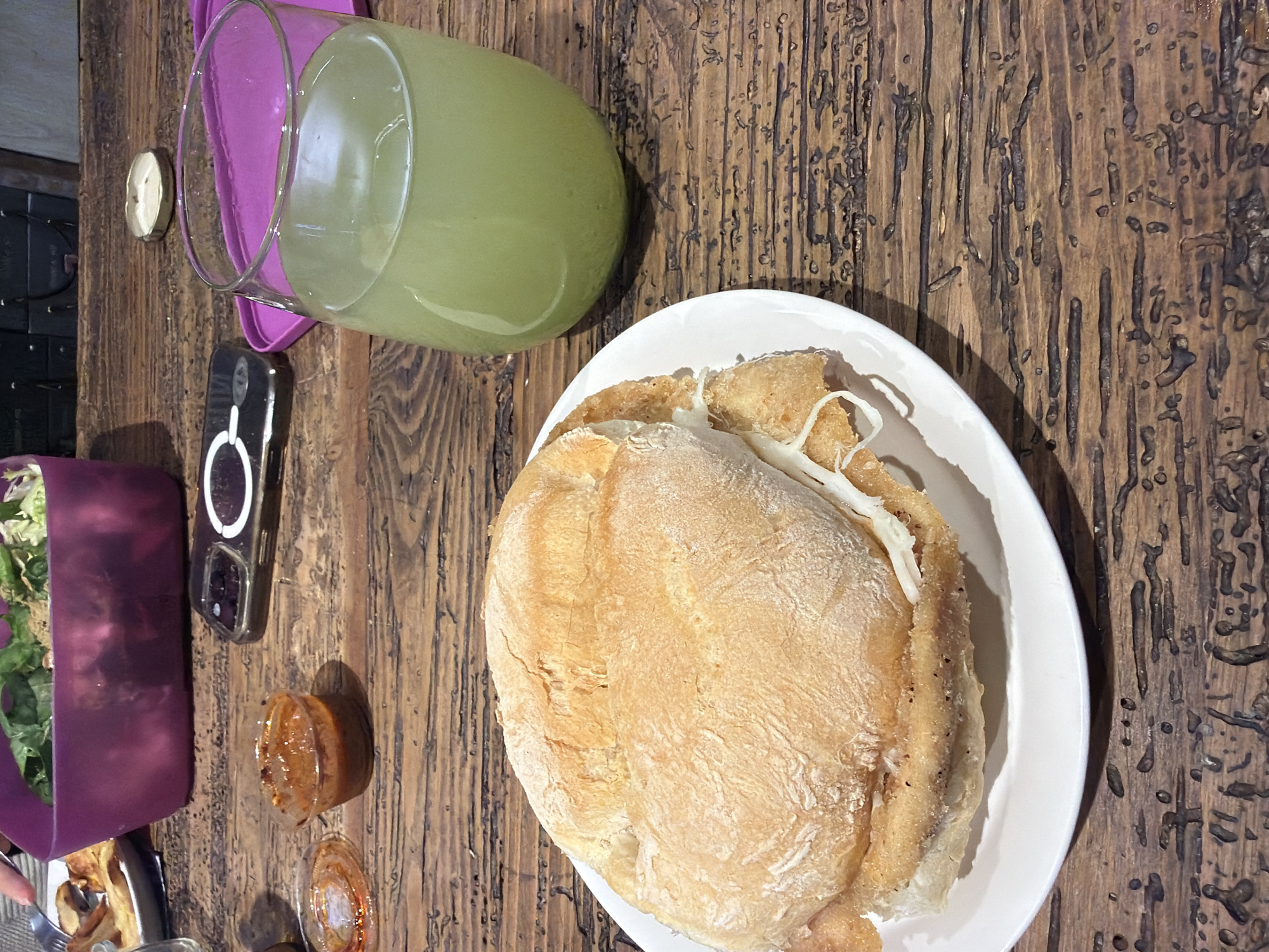 The image shows a table in what appears to be a restaurant setting, likely in Puebla, Mexico, based on the presence of a cemita, a type of sandwich typical of the region. On a wooden table is a white plate with a large cemita sandwich. Next to the plate is a tall glass filled with a green beverage, potentially agua fresca or juice. Nearby are a small container with an orange sauce (possibly a salsa), a cell phone, and a purple container filled with salad. A small condiment cup, possibly containing butter or sauce, sits to the side of the glass. The table has a textured, possibly rustic or distressed, finish.