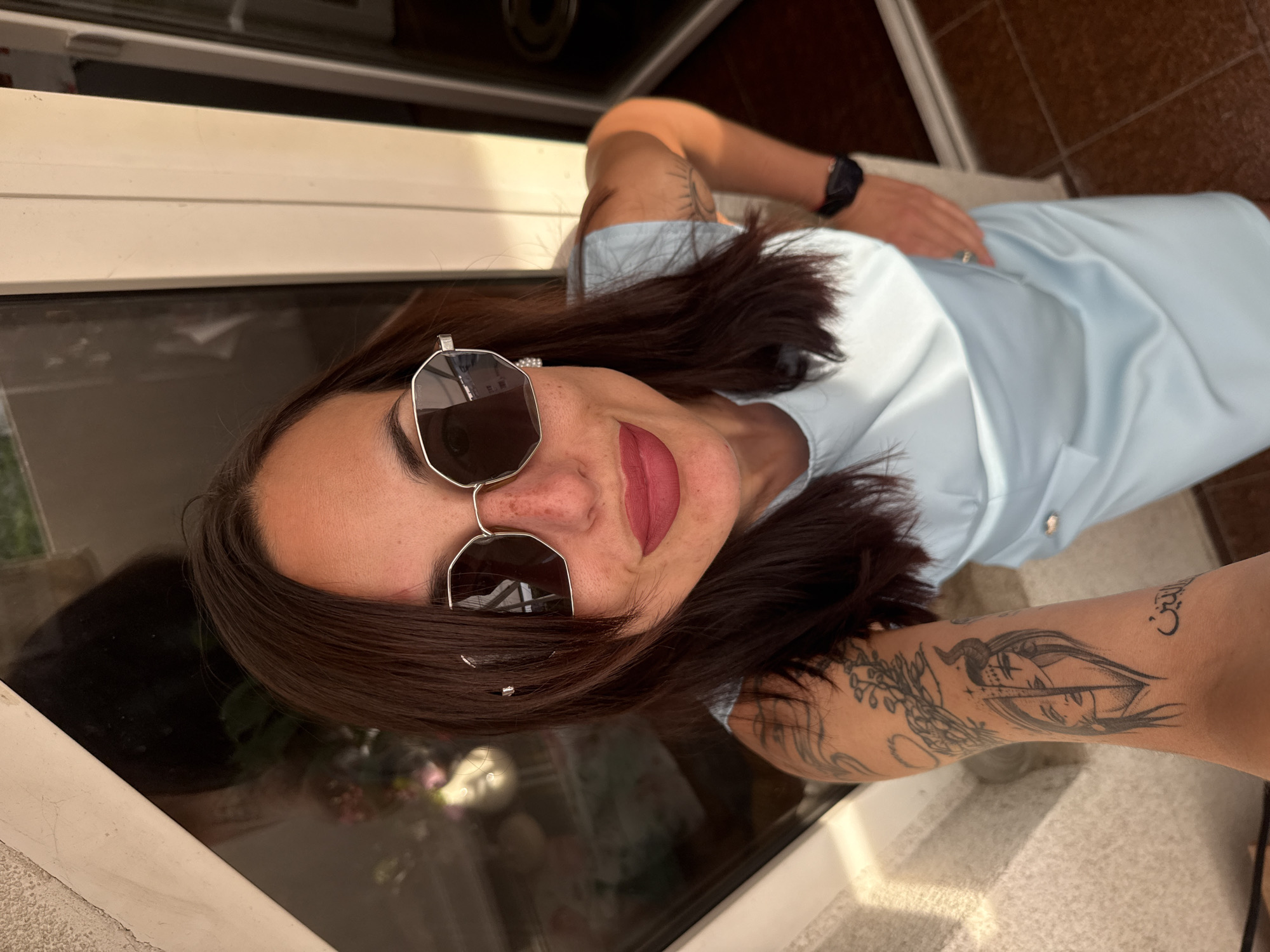 A woman wearing a light blue shirt and sunglasses is seen in a close-up shot. She is positioned near a window. Visible on her arms are tattoos. The surroundings suggest an interior setting, possibly in Pleven, Bulgaria.