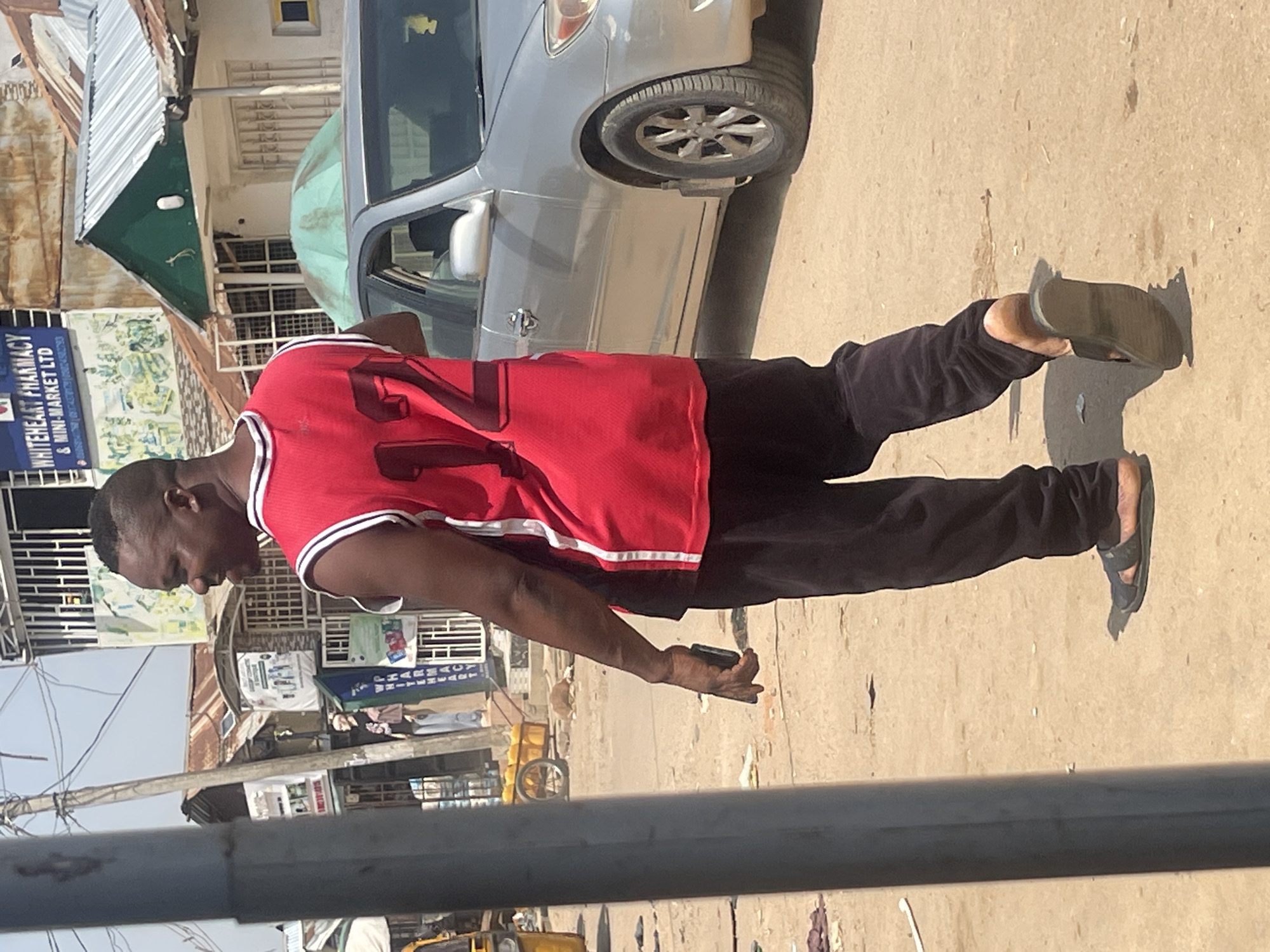 A man walks down a dirt street in Abuja, Nigeria, wearing a red basketball jersey with the number 12, black pants, and black sandals. He is holding a dark-colored smartphone in his right hand. Behind him, a silver sedan is parked, and beyond that, a building with "WHITEHEART PHARMACY & MINI-MARKET LTD" signage is visible. The environment appears to be an outdoor market or street with various stalls and structures. The sky is clear and bright, indicating a sunny day.