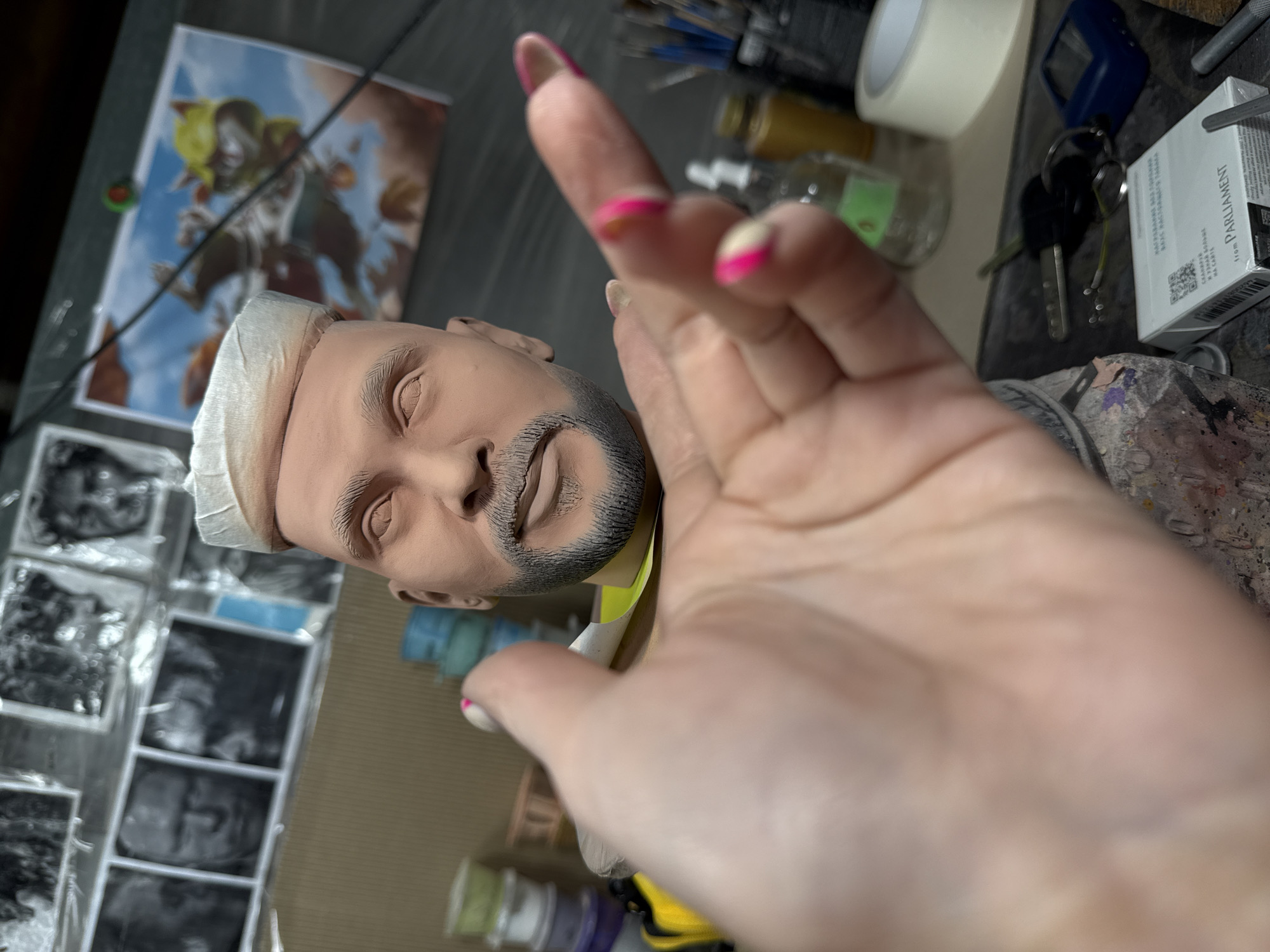 The image shows a person's hand with pink-tipped nails partially obscuring a clay sculpted head of a man with a trimmed beard and light-colored hair. The head is positioned near a framed print of what appears to be fantasy art. To the left, there's a strip of black and white photos. The background includes various items like tape, brushes, small bottles, keys, a blue electronic device, and a pack of Parliament cigarettes. No definitive evidence places the scene in Chaikovsky, Russia.