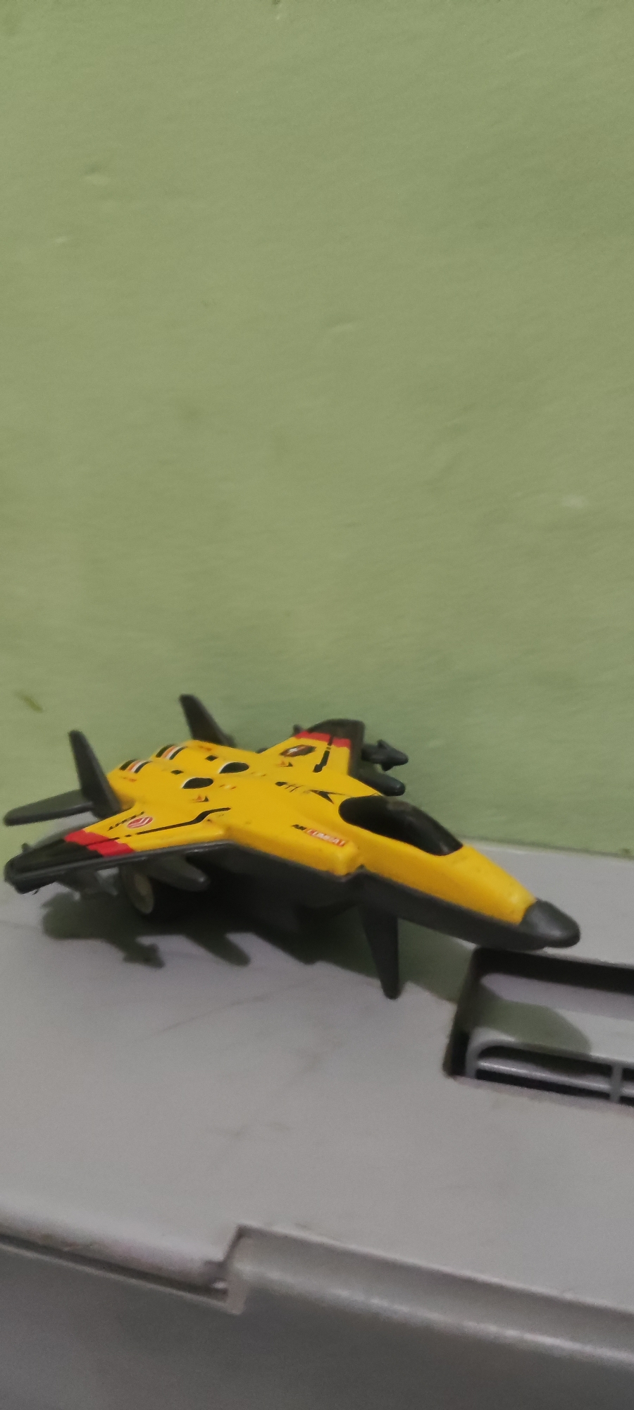 A yellow and black toy jet fighter with red accents is positioned on a grey surface in front of a light green wall. The jet has visible markings including what appears to be "AIR COMBAT" on its side. The surface it rests on has a visible edge or lip. The image is taken from a slightly elevated angle, with the jet angled towards the right. The background is a plain, slightly textured green wall. The scene appears to be indoors, likely in a residential setting in Gebang, Indonesia.