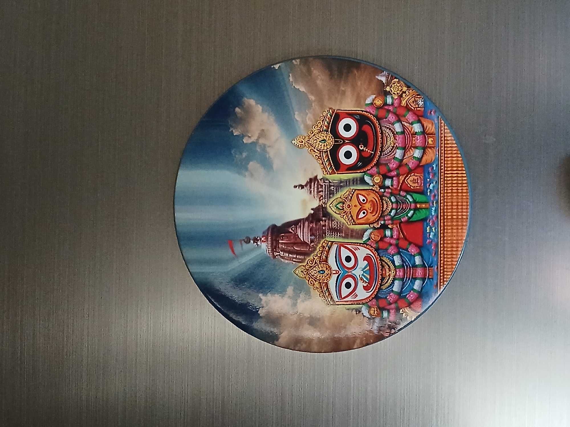 The image displays a circular, glossy decorative item, likely a plate or magnet, affixed to a textured, brushed metallic surface, possibly a refrigerator door, in Bengaluru, India.
The item features a vibrant illustration of the three principal deities of the Jagannath Temple: Lord Jagannath, Subhadra, and Lord Balabhadra. Lord Jagannath, with a black face and large white eyes, is positioned at the top of the triad, adorned with golden headwear and colorful garlands. Below him, Subhadra is depicted with a yellow-orange face and wide eyes, wearing green and floral adornments. At the bottom is Lord Balabhadra, recognizable by his white face with distinctive red and blue markings around his large white eyes, also richly decorated with golden ornaments and flower garlands.
In the background of the illustration, the grand shikhara (tower) of the Jagannath Temple in Puri is visible, topped with a red flag. The sky behind the deities and temple transitions from a bright blue with white clouds on the left to warmer, golden hues on the right, suggesting either sunrise or sunset, with rays of light emanating through the clouds. The overall scene conveys a sense of divine presence and spiritual reverence, characteristic of Hindu iconography. No specific activity is depicted other than the presentation of these revered figures. There is no visible text on the item. The metallic background features vertical grain lines and varies in brightness.