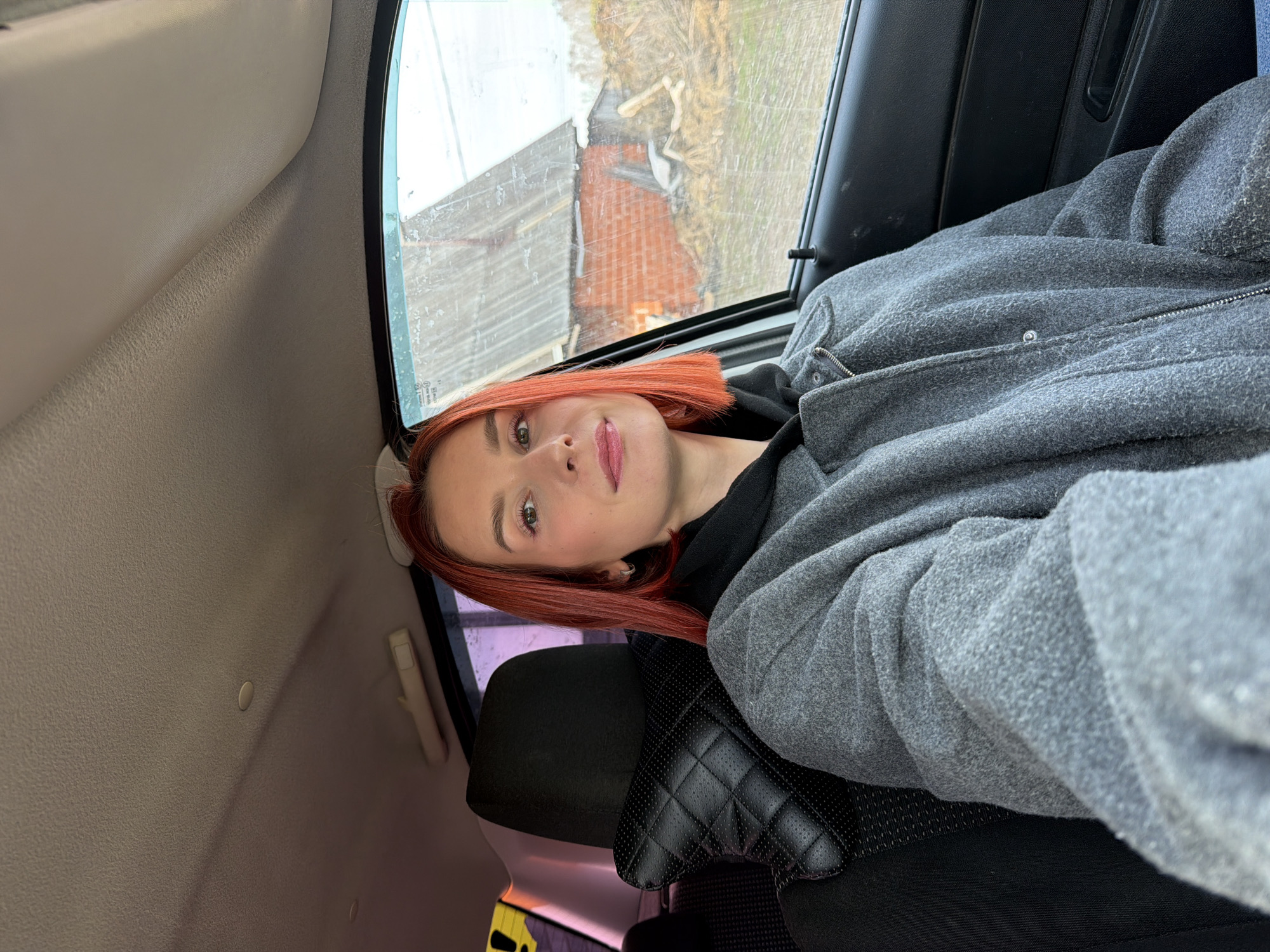 A woman with shoulder-length, auburn-colored hair is seated in the back seat of a car. She is wearing a gray, zippered, long-sleeved jacket over a black hooded shirt. The car seat is black leather with a quilted pattern. Outside the car window, a dilapidated building with a brick facade and a corrugated metal roof is visible, as are bare branches of trees. The location is potentially Chaikovsky, Russia, as per the prompt.