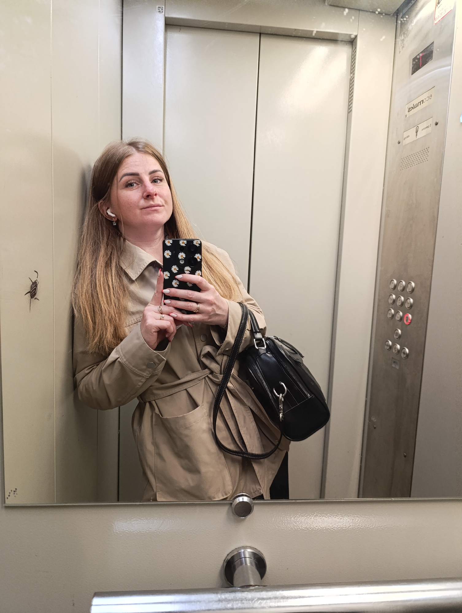 A young woman stands in an elevator, taking a mirror selfie. She has long, blonde hair and is wearing a tan trench coat and holding a black purse. Her phone, with a daisy-patterned case, is visible as she takes the photo. The elevator interior is utilitarian, with brushed metal panels and buttons. A small, insect-like sticker is visible on the wall to the left of the woman. The overall impression is mundane, capturing a fleeting moment in an everyday urban setting.