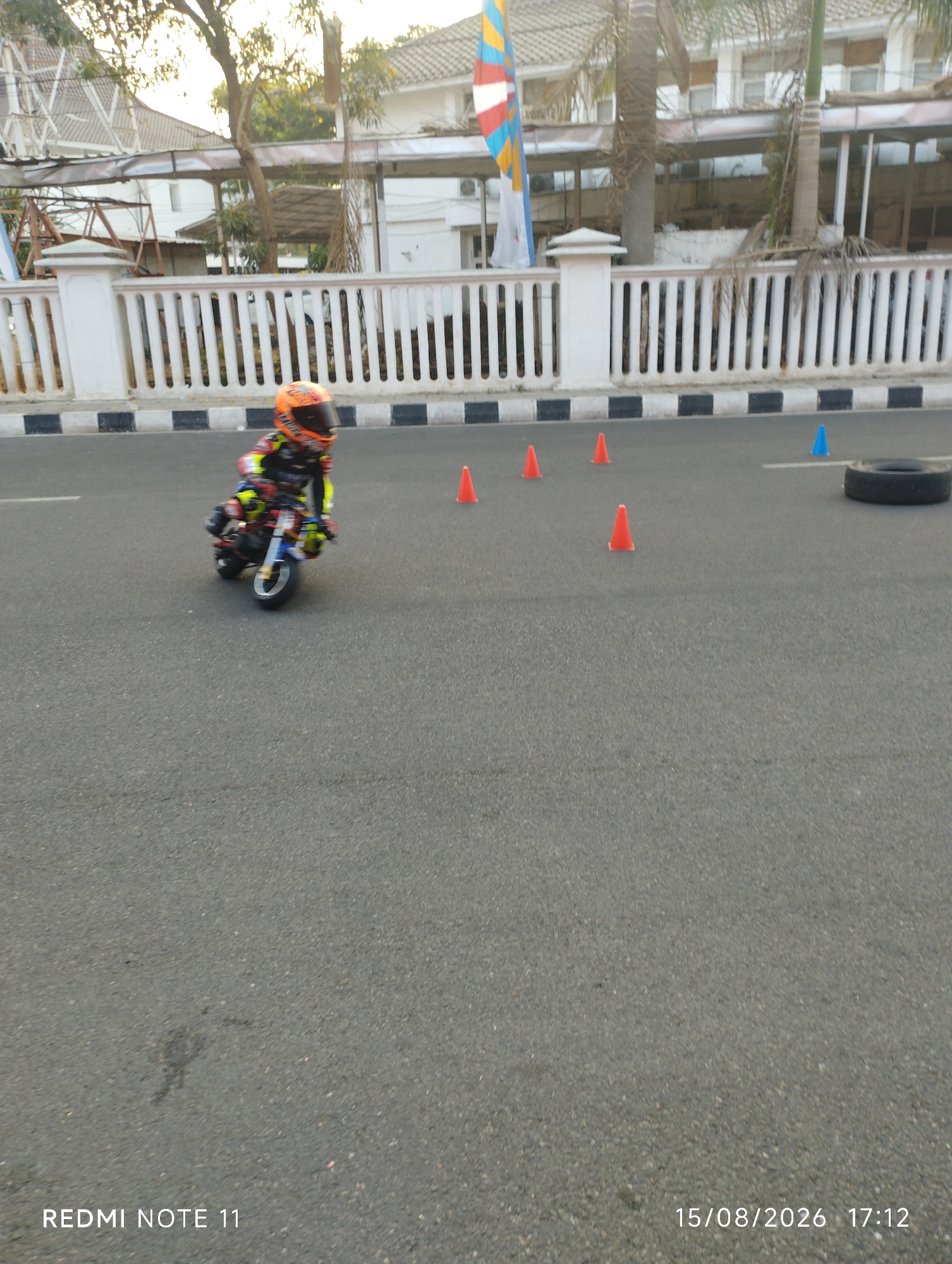 A child in a full racing suit and helmet is riding a mini motorcycle on an asphalt road. The child is wearing an orange helmet with black and yellow accents, and a black and yellow racing suit with red and blue details. The motorcycle is blue and black with white wheels. The road is lined with a white fence with black and white stripes at the bottom. Orange and blue traffic cones are placed on the road, suggesting a practice or training course. In the background, there is a building with a white facade and a tiled roof, surrounded by trees. The scene is outdoors, likely in a public space or a dedicated training facility, given the presence of the cones and the organized layout. The timestamp on the image indicates the date is August 15, 2026, at 5:12 PM. The location is identified as Karawang, Indonesia.