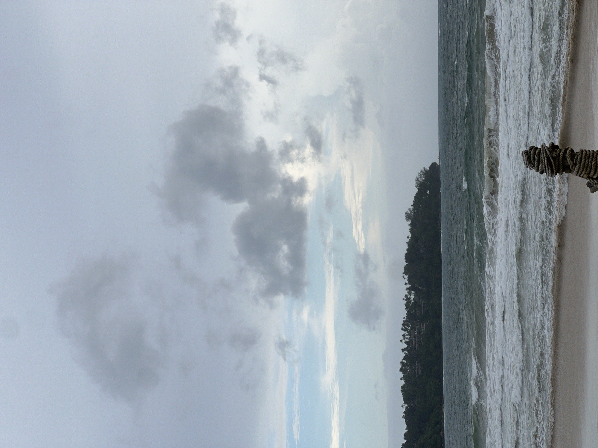 A landscape photograph, oriented vertically, depicts a beach scene under an overcast sky. The left half of the image is dominated by various shades of gray clouds, interspersed with patches of lighter sky. The right half shows the ocean meeting the beach. Waves crash onto the shore, with white foam contrasting against the gray water. A dark green, tree-covered land mass extends into the ocean. In the foreground on the right, a thick, knotted rope is visible.