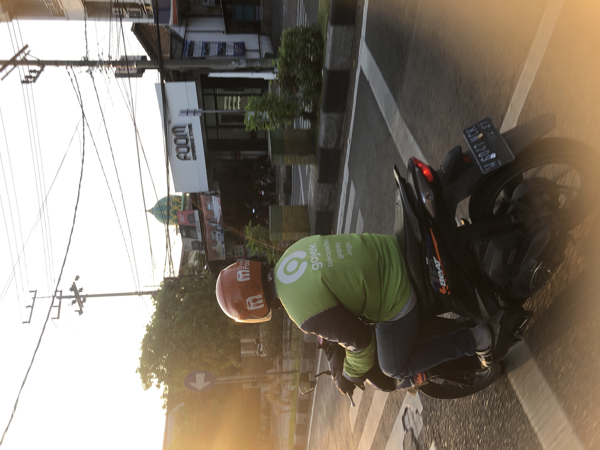 A man wearing a bright green Gojek jacket and an orange helmet is riding a black motorcycle. He is on a road in what appears to be the city of Sidoarjo, Indonesia. The sun is setting, casting a warm glow on the scene. In the background, there are buildings, a traffic light, and power lines. The motorcycle has a license plate that reads "W 6047 VCK". The Gojek driver is wearing blue jeans and sandals. The motorcycle is a Honda Beat.