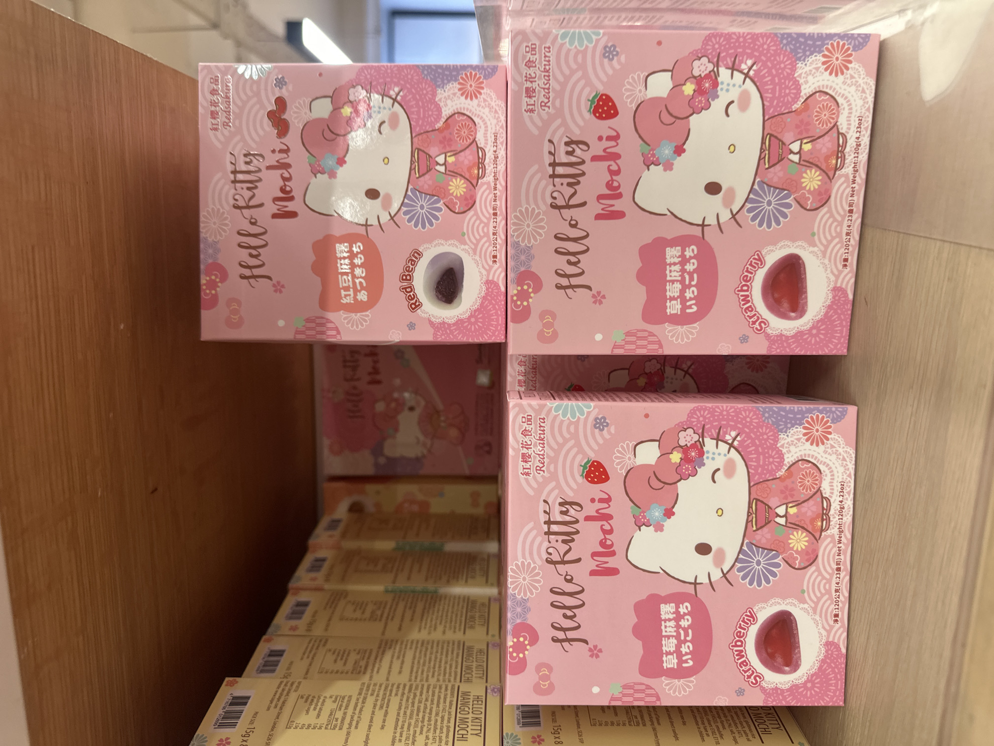 The image shows a shelf in an indoor retail environment stocked with boxes of "Hello Kitty Mochi" candy. The location is not discernible from the image alone, and therefore, London, United Kingdom cannot be confirmed. The topmost boxes are predominantly pink and feature images of Hello Kitty in a kimono, with labels indicating flavors like "Red Bean" and "Strawberry." Below, more boxes of what appear to be "Hello Kitty Mango Mochi" are stacked. Nutrition information and manufacturer details are printed on the sides of the Mango Mochi boxes. The overall setting suggests a store aisle.