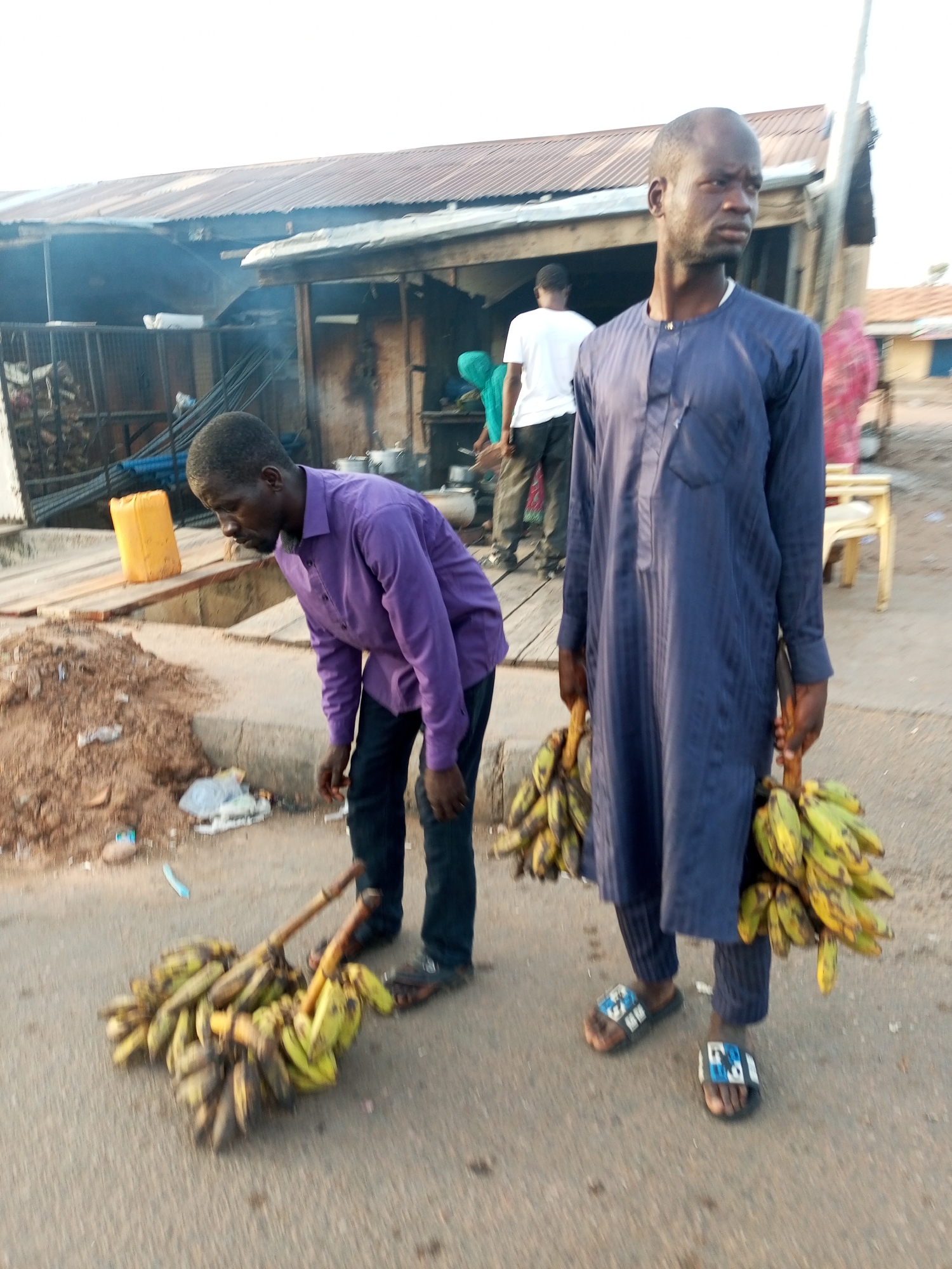 Two men stand on a paved street, likely in Gombe, Nigeria. The man on the left wears a purple long-sleeved shirt and dark jeans, bending over a bundle of plantains resting on the ground with two sticks running through the middle. The man on the right is wearing a blue long-sleeved tunic and pants, holding another bunch of plantains. In the background, there is a corrugated iron structure with people near what appears to be cooking equipment. A yellow jerrycan sits nearby. Other buildings are visible.