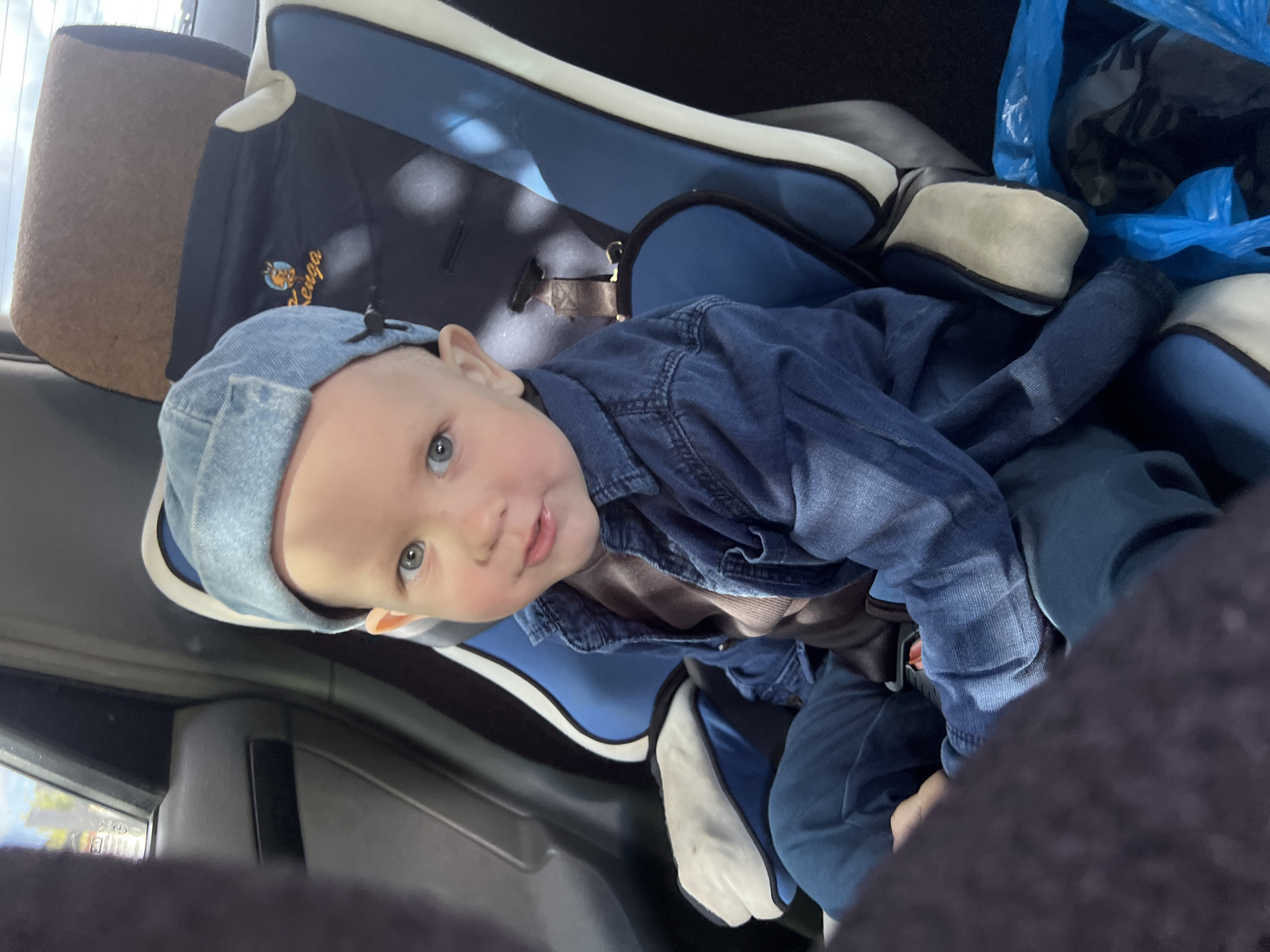 A close-up, high-angle shot depicts a baby wearing a light blue denim cap and jacket, sitting in a car seat. The car seat has a blue and white trim. Parts of the car's interior, including the seat and door panel, are visible. A blue plastic bag sits next to the car seat. The location is not discernible from the image.
