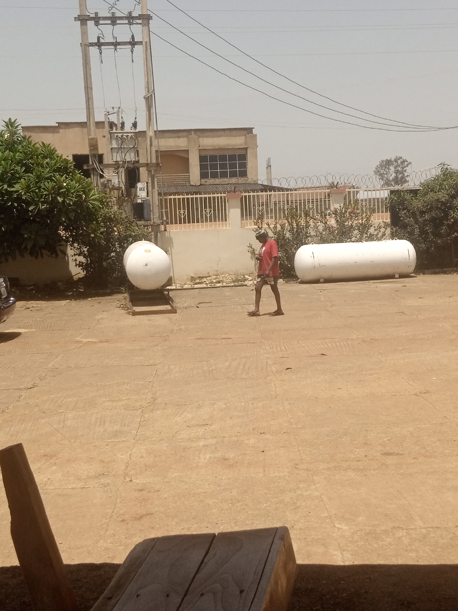 A person in a red t-shirt and shorts walks across a concrete surface in front of two large white cylindrical tanks. To the left of the person, one tank is upright, while the other, to the right, lies horizontally. In the background, a building with a barred window is visible, along with a white fence topped with barbed wire. A utility pole with electrical wires stands tall in the left background. Trees and bushes are scattered throughout the scene, and the ground is a dusty, light brown. The sky is hazy, suggesting it might be a warm, possibly dry, day. The overall impression is an outdoor industrial or storage area. No visible text is present in the image.
