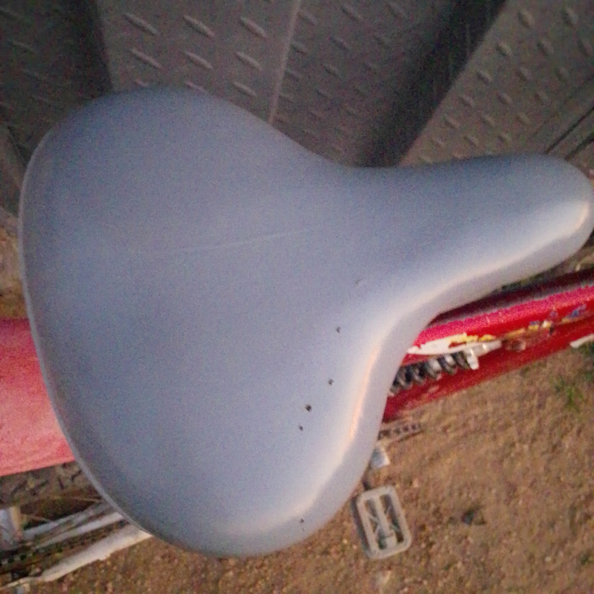 The image shows a close-up of a gray bicycle seat positioned on top of a red bicycle frame. The seat is smooth and slightly curved, with a matte finish. There are a few small, dark specks on the surface of the seat, likely indicating minor imperfections or dirt. The bicycle frame appears to be metal, painted red, and has some visible wear and tear, including scuffs and possibly some rust. The background is blurred, but suggests an outdoor environment with a textured surface, possibly dirt or gravel, and some greenery. The lighting suggests it is daytime, with a warm, golden hue. The location in Wuro Allahamdu, Nigeria, is not explicitly depicted or suggested by any elements within the image itself; the scene is a generic view of a bicycle.